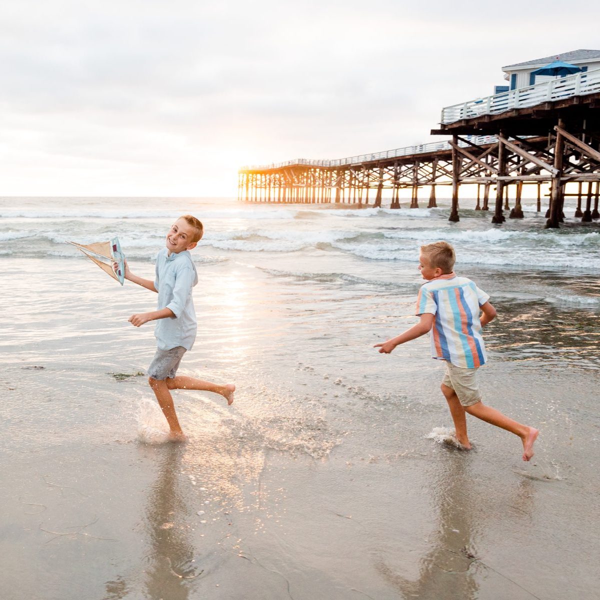 Two brothers running and splashing along the shoreline near a wooden pier