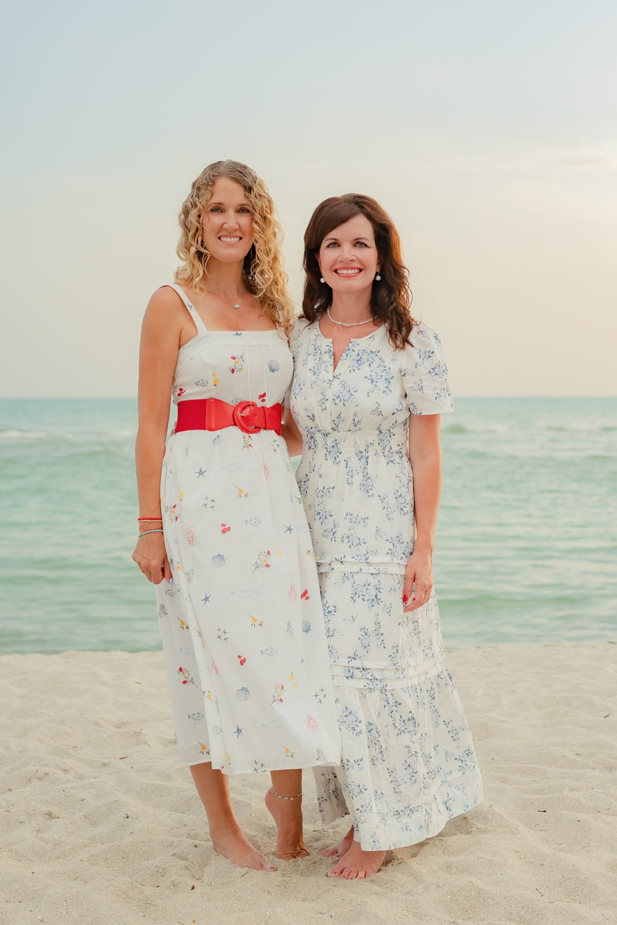 Mother and daughter in white dresses smile on the sand