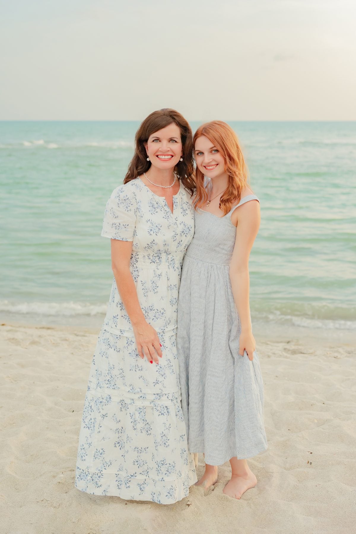 Mother and daughter in flowing dresses smile by the sea