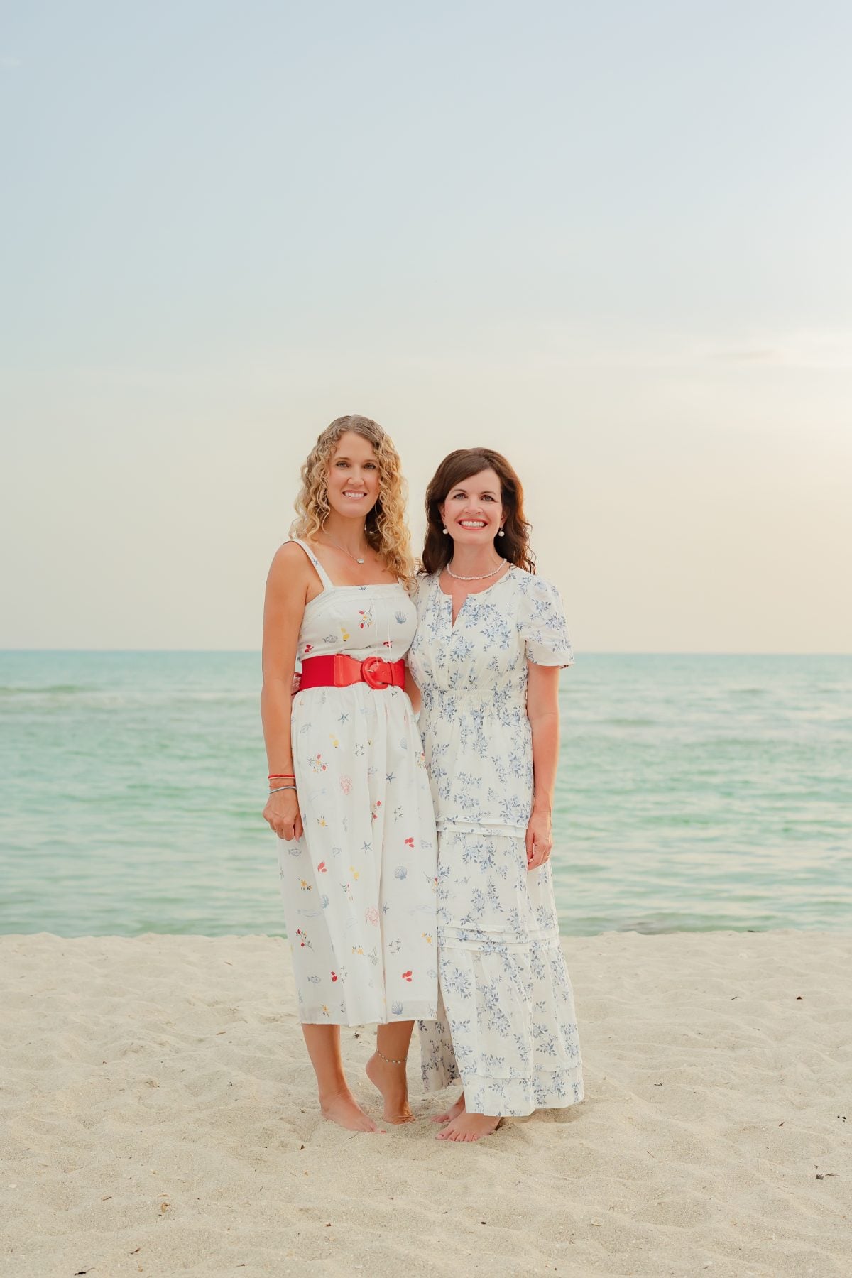 Two women in matching white dresses embrace beachside