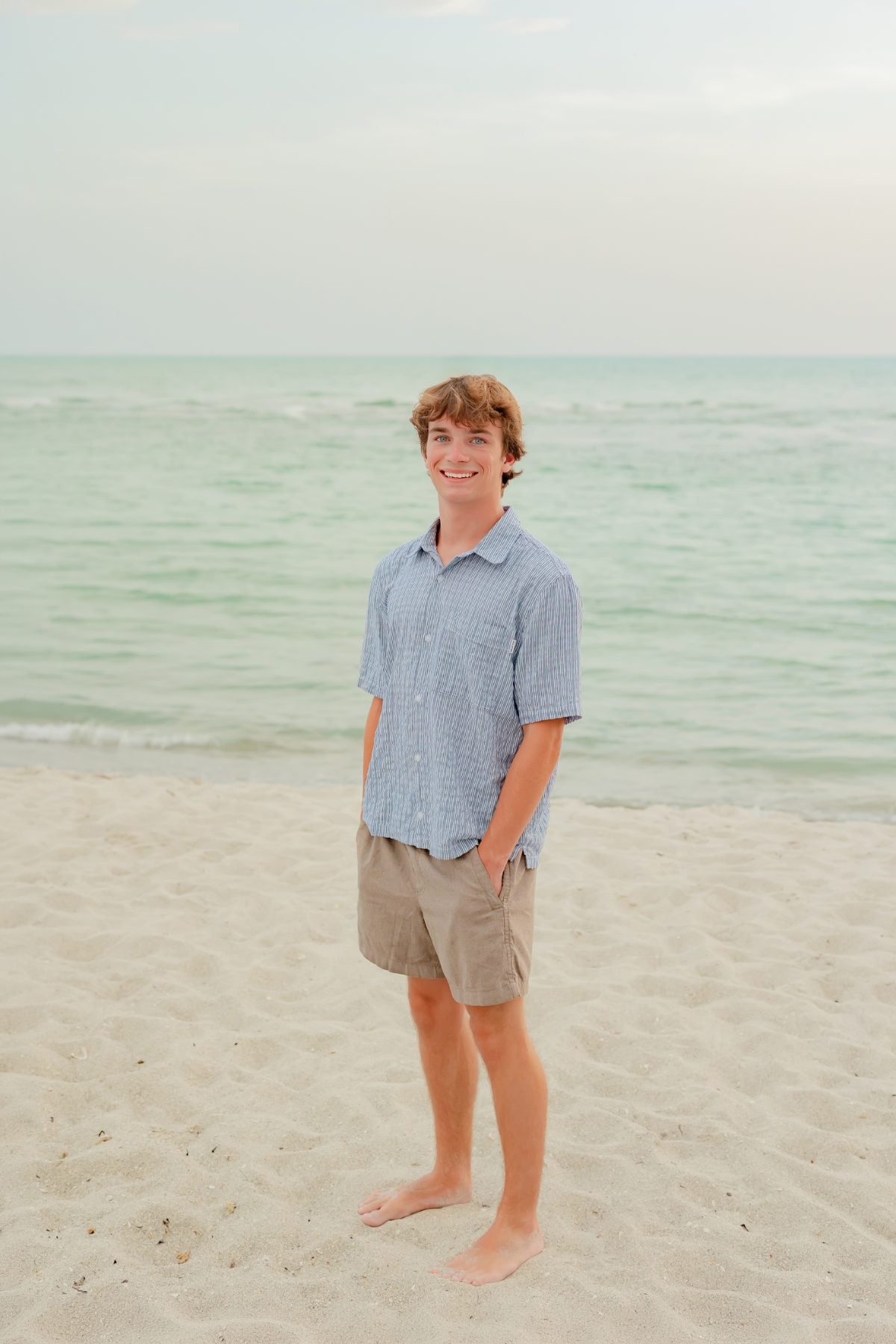 Teen boy in blue shirt stands barefoot on sandy beach
