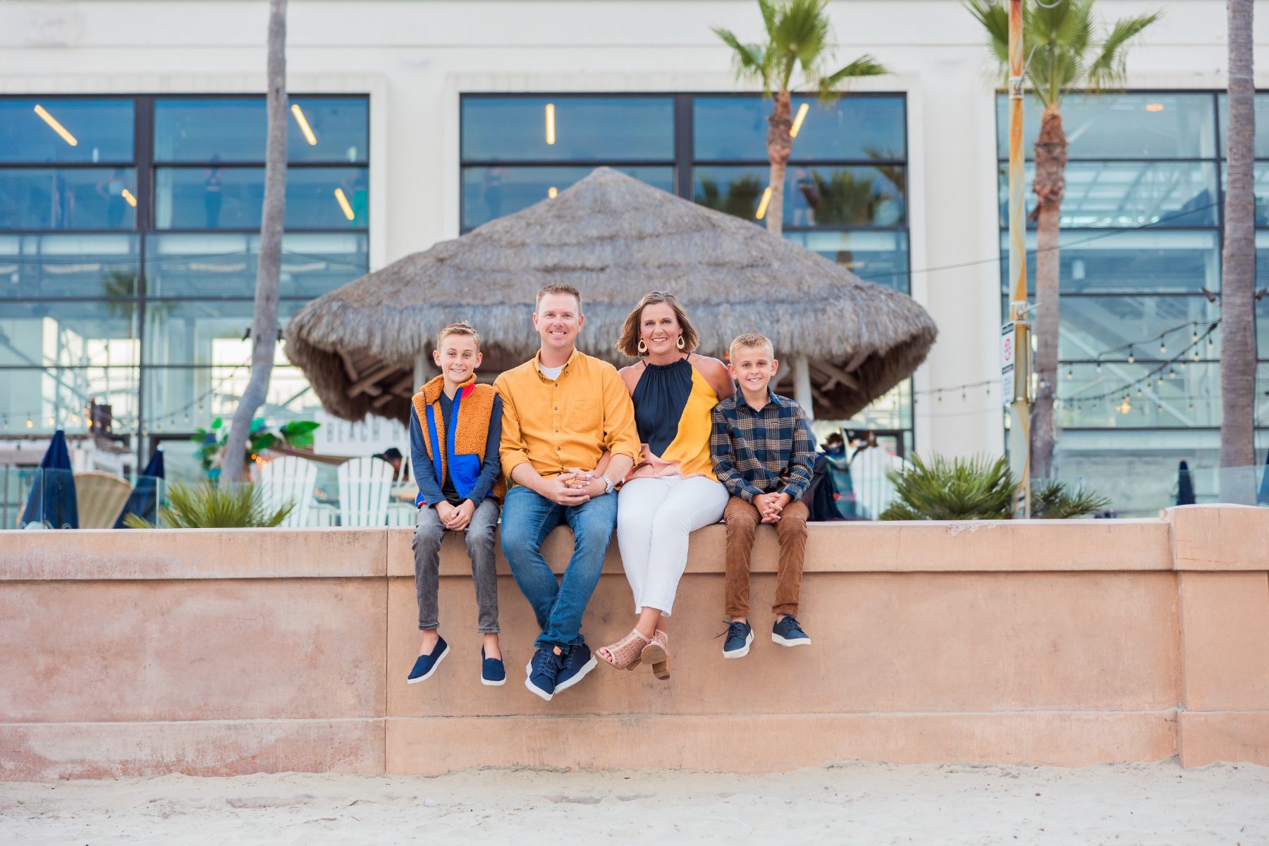 Family of four sits together on a wall near palm trees and a thatched umbrella