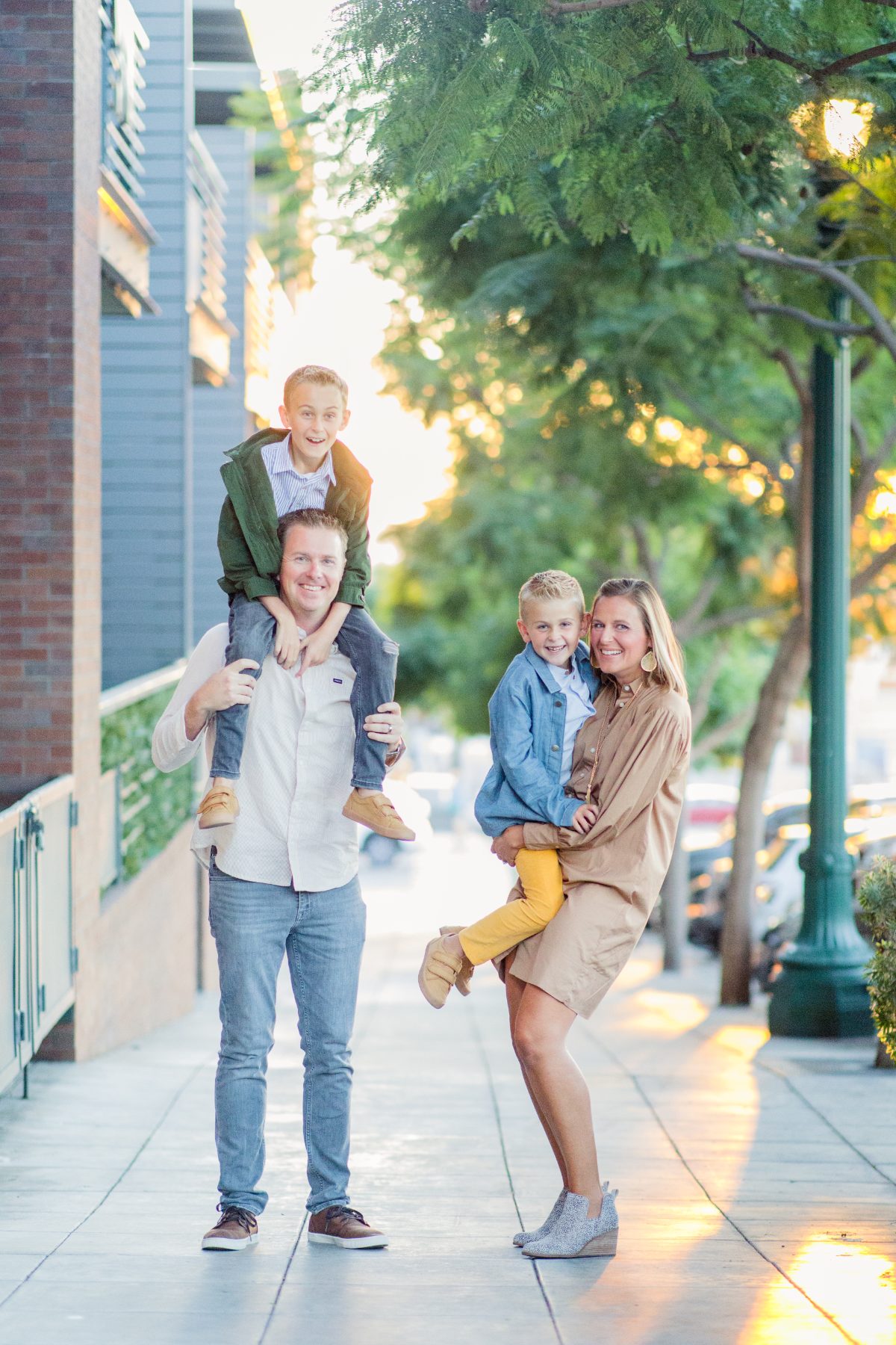 Dad carries son on shoulders while mom holds other son walking downtown sidewalk