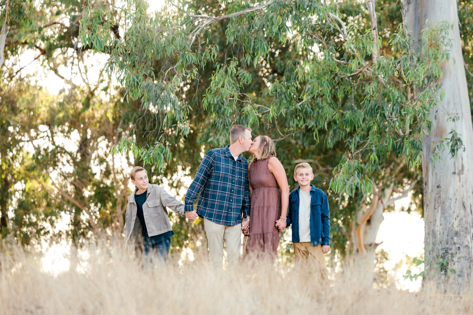 Dad kisses mom while sons hold hands walking through tall grass