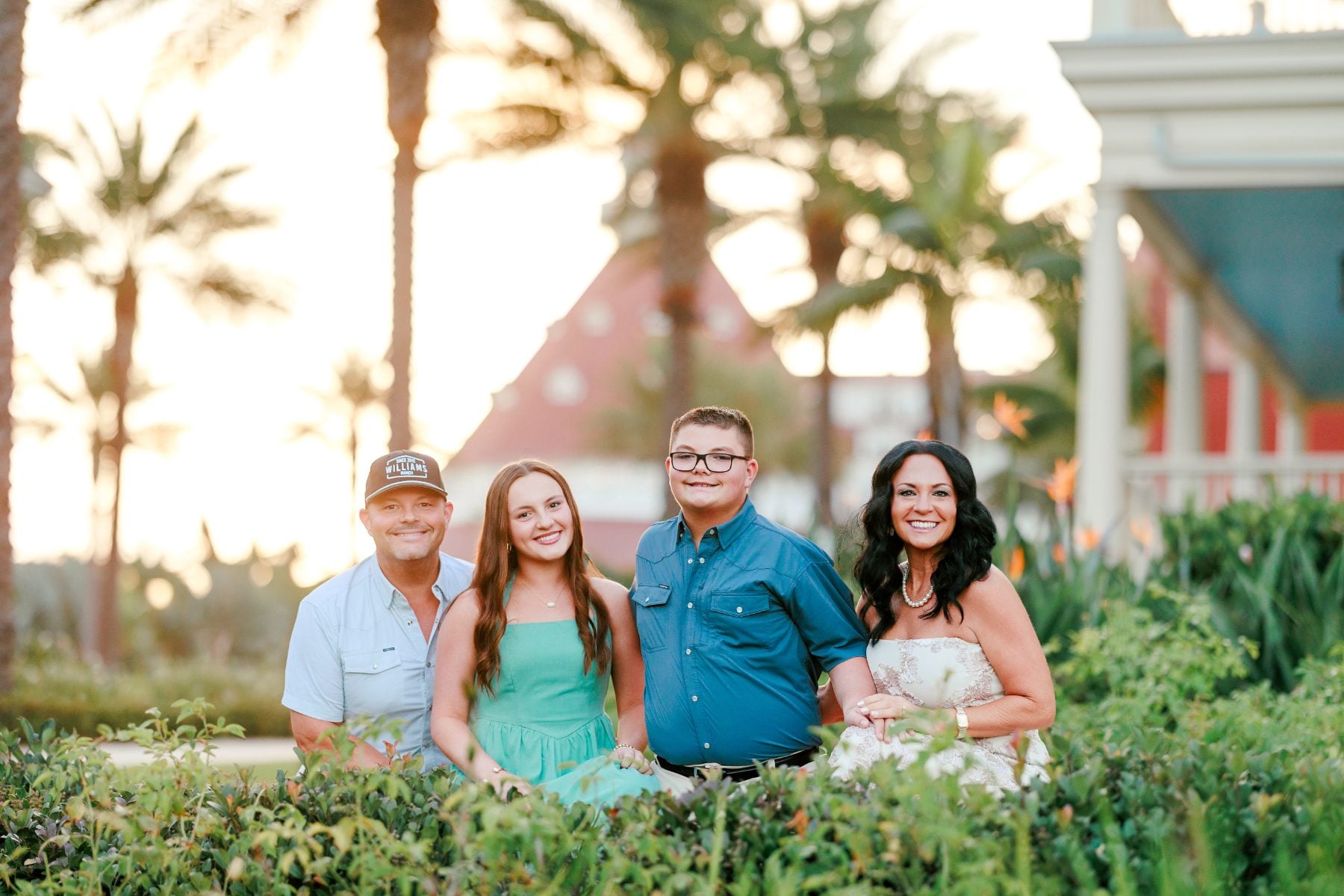 Extended family of four sits together in garden, Hotel del Coronado behind them