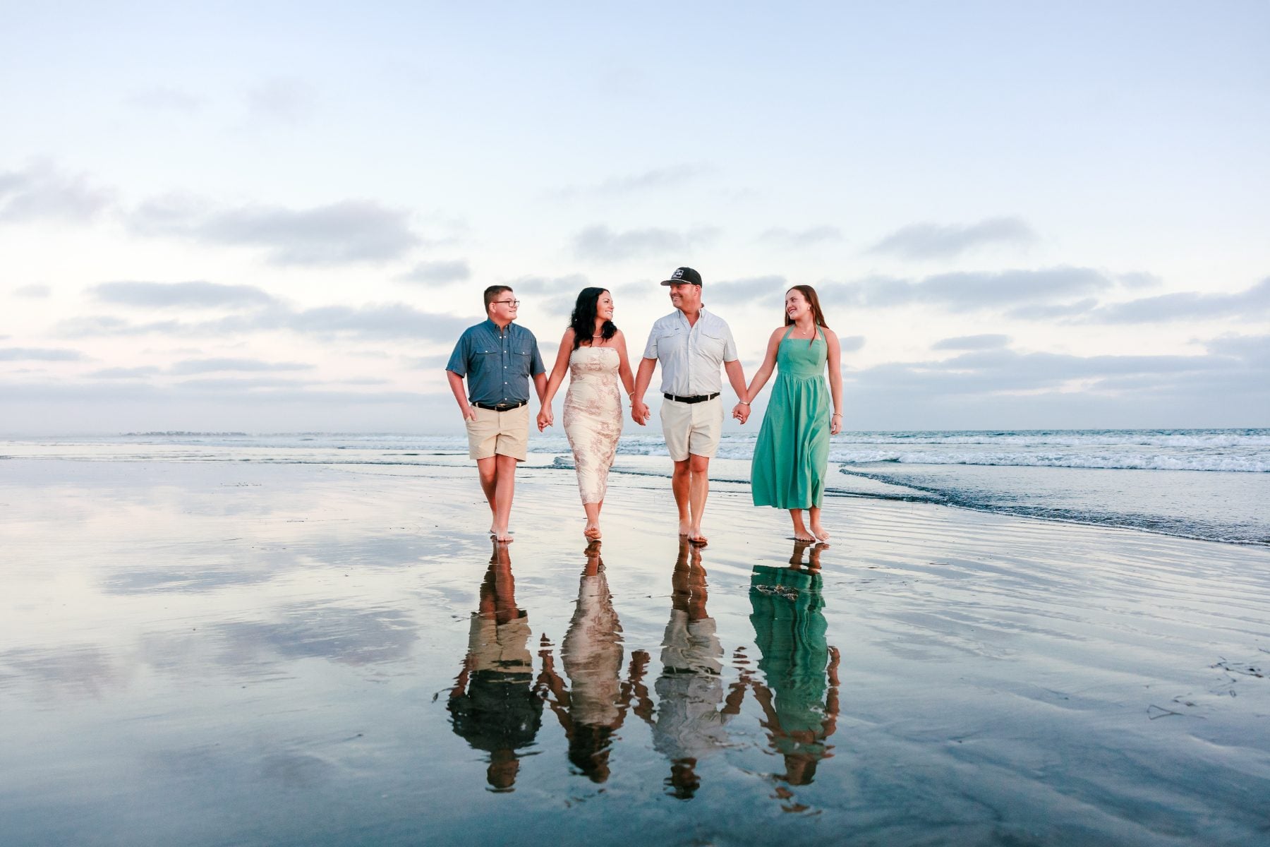 Family of four holds hands walking on wet sand reflections, Coronado Beach