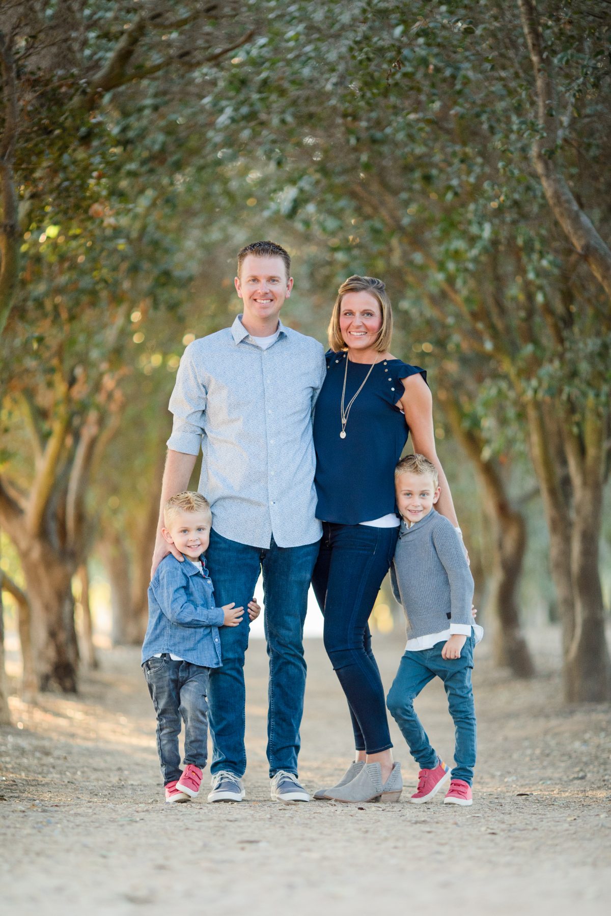 Family of four stands smiling under shaded tree-lined orchard path