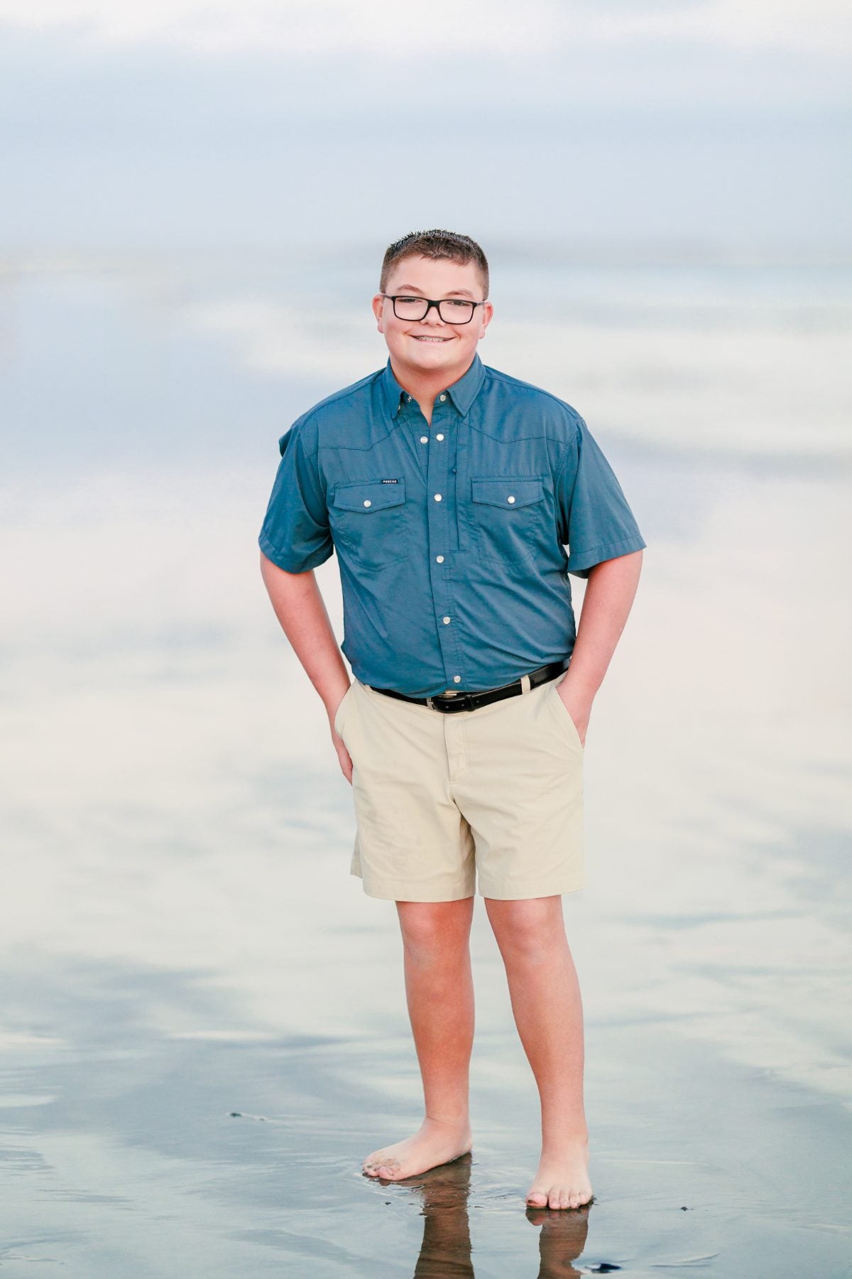 Teen boy stands barefoot on the beach in blue shirt, khaki shorts