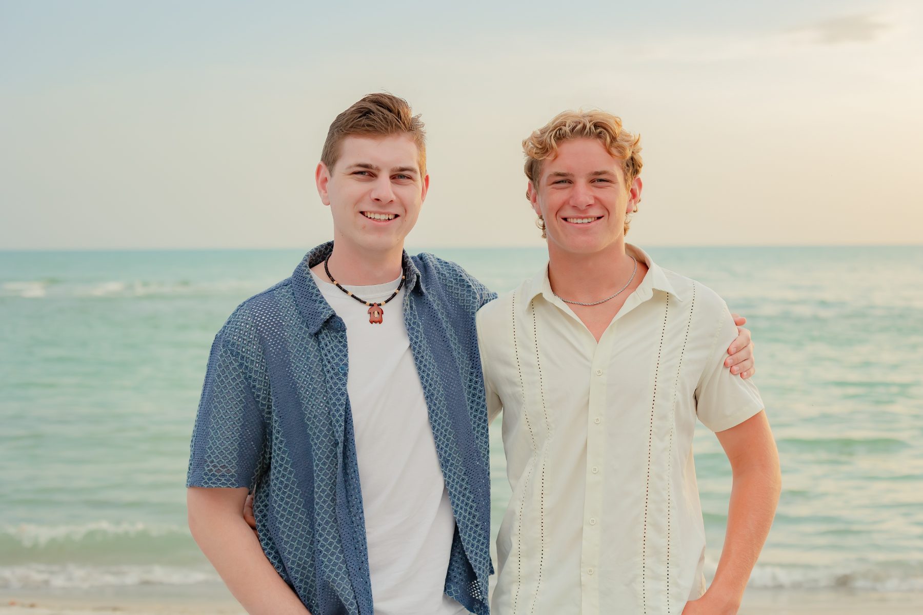 Two brothers in patterned shirts pose arm in arm on the beach