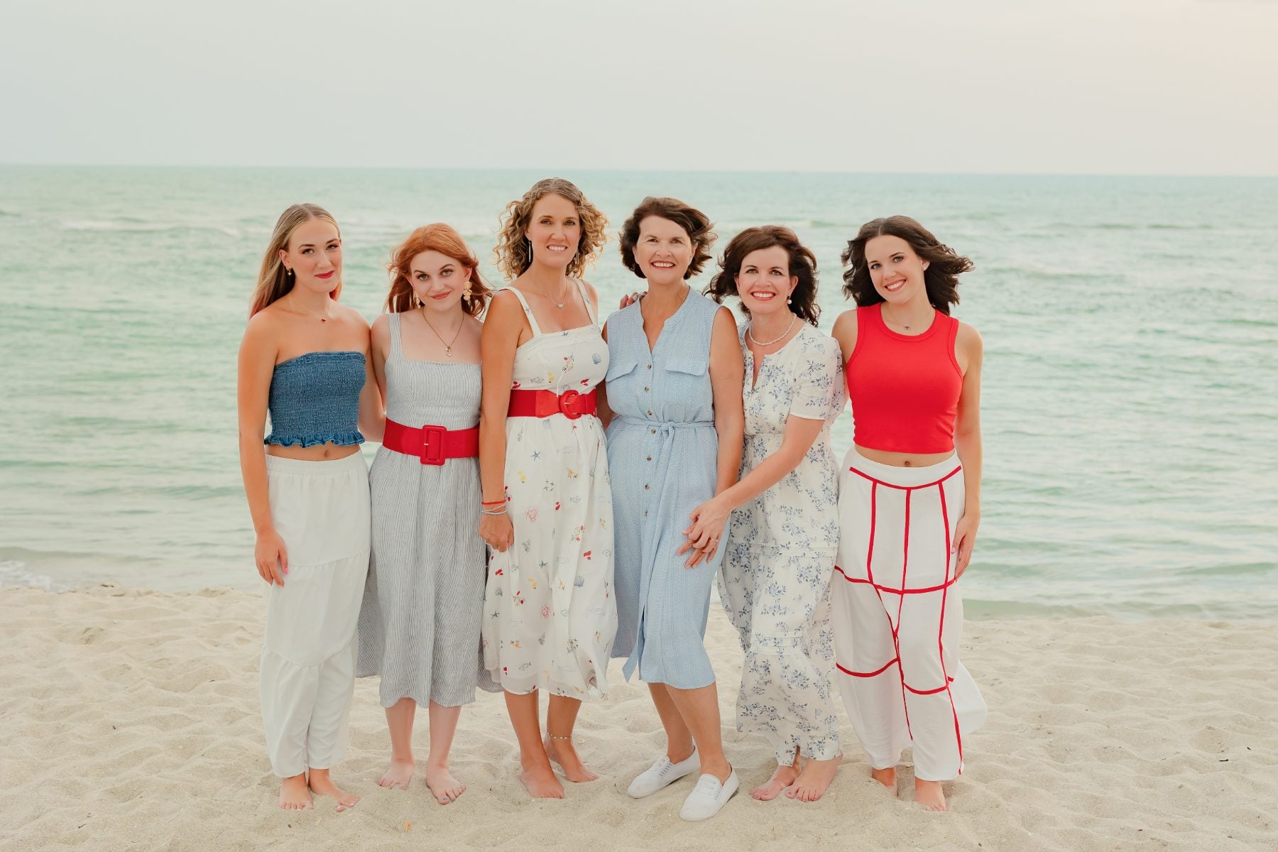 Six women in coordinated outfits pose along the shoreline