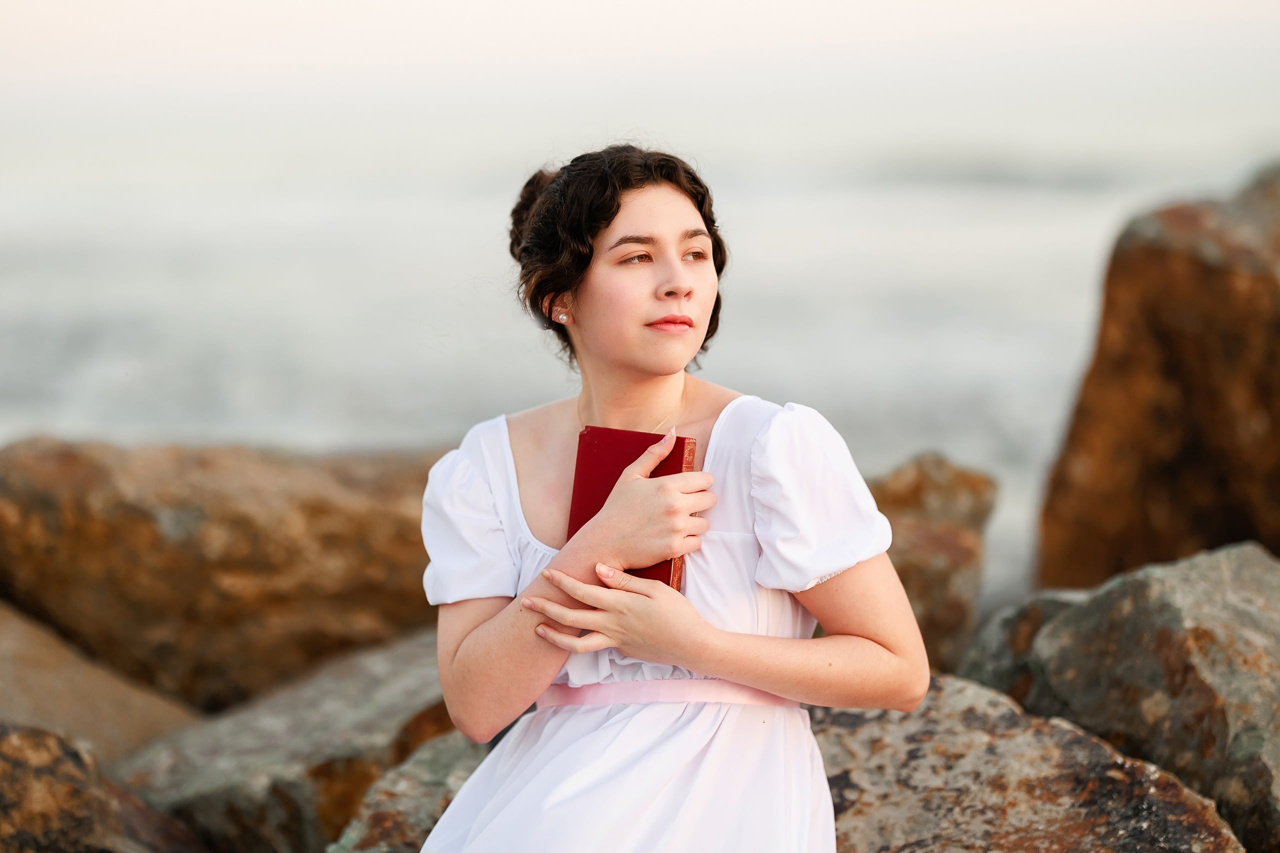 Amanda Fagan’s Love, Jane — 20 Woman in white dress holding red journal standing on rocks overlooking ocean