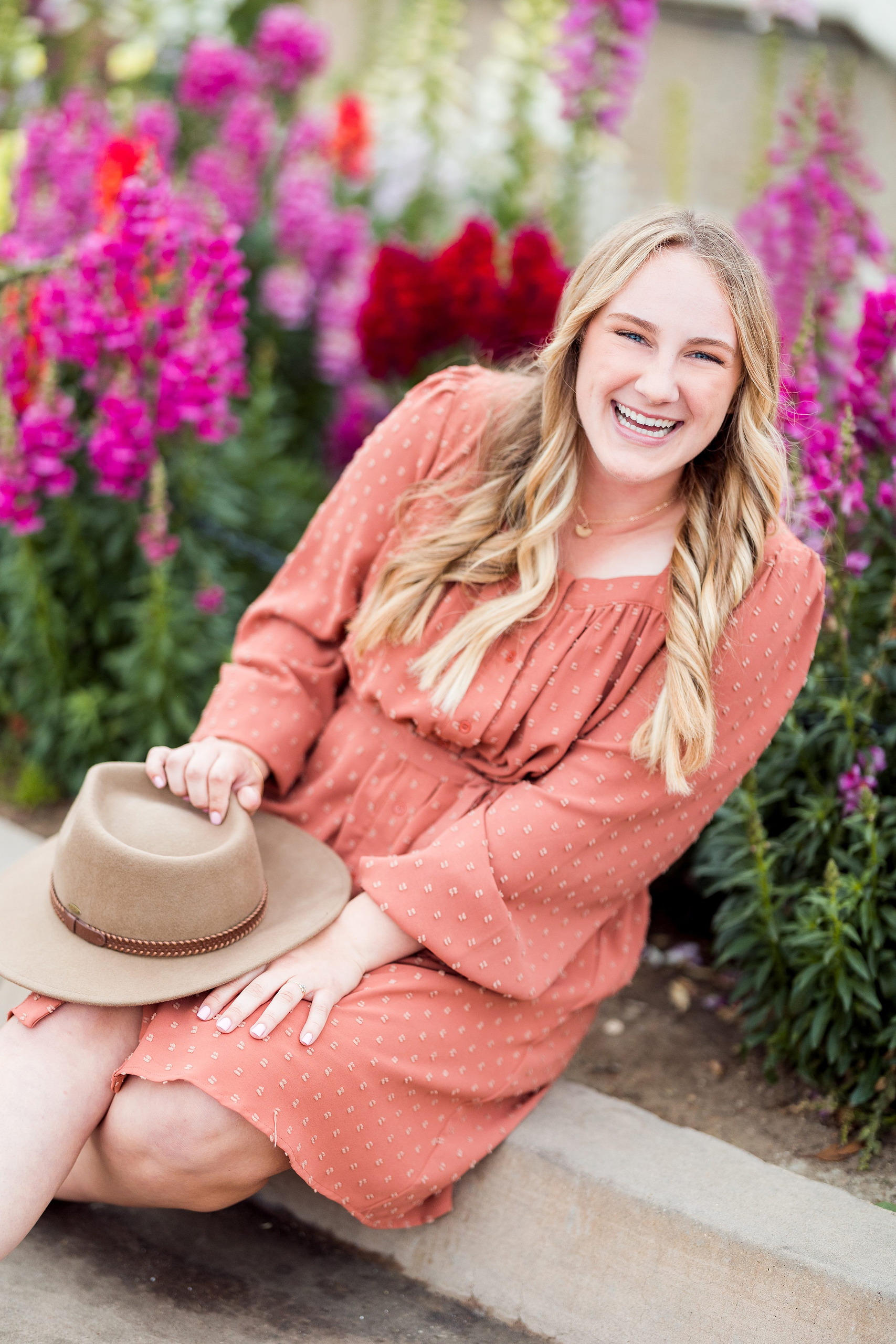 Balboa Park Senior Photography Session — 02 Senior kneeling in coral dress holding tan hat with pink flowers behind