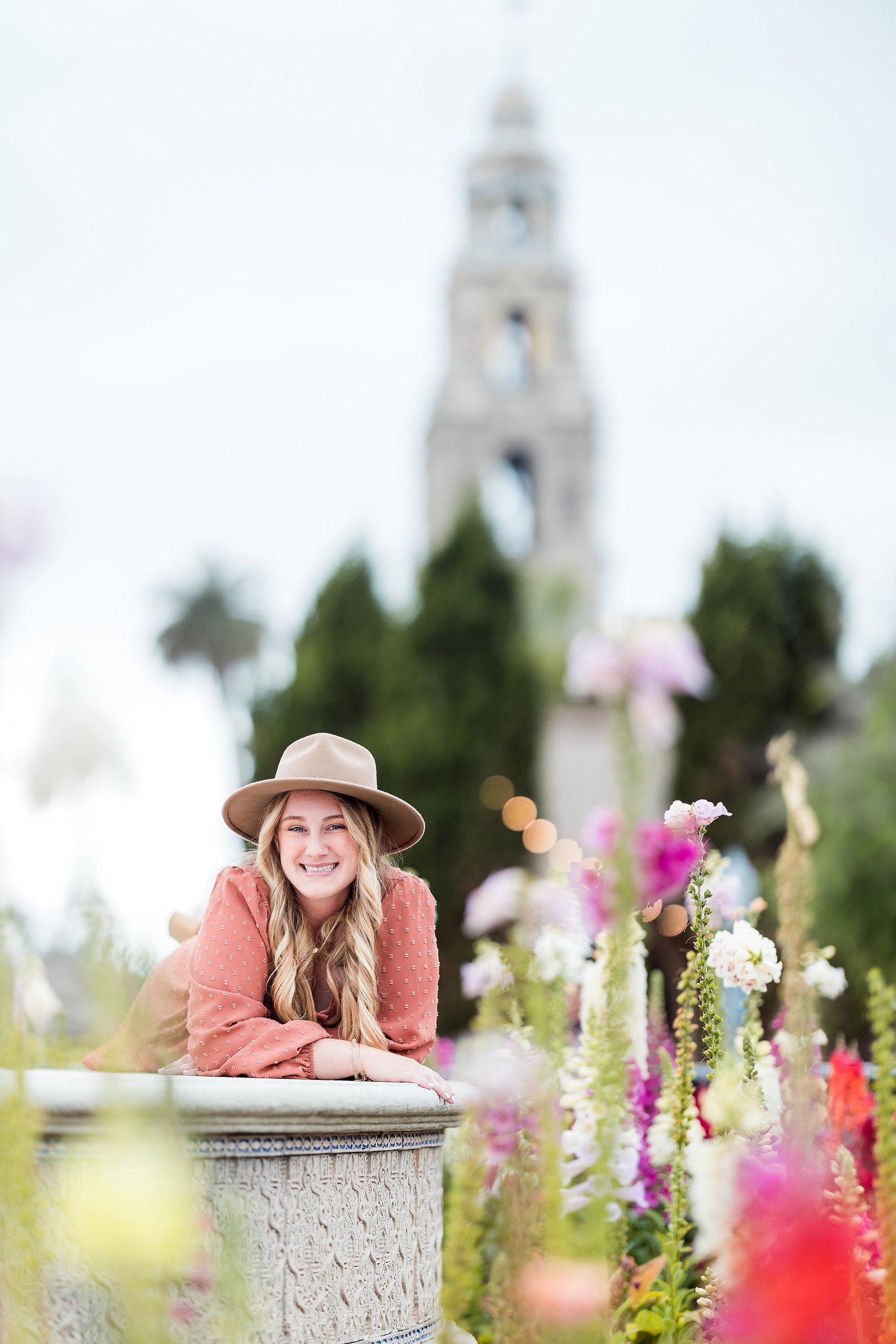Balboa Park Senior Photography Session — 04 Senior leaning on stone ledge with flowering garden and bell tower in background
