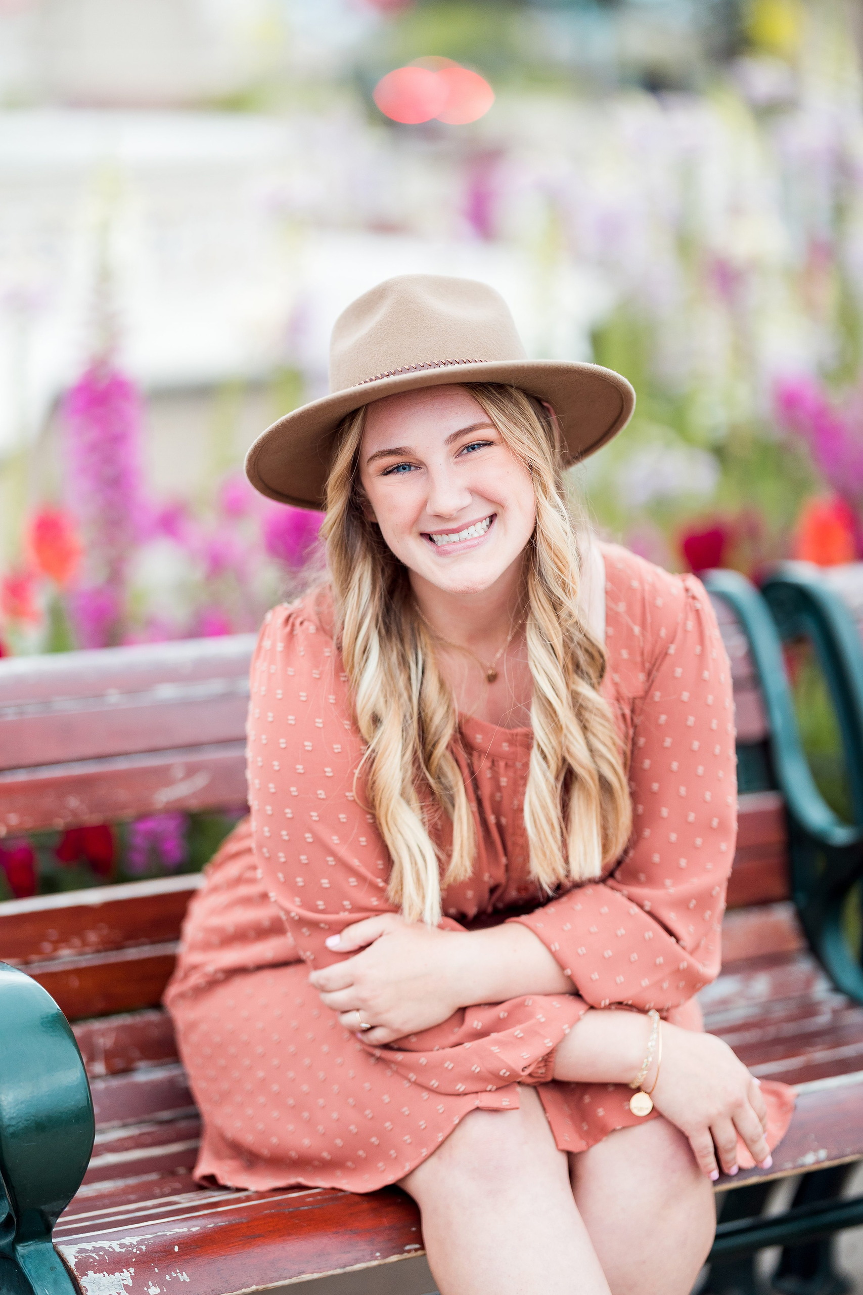 Balboa Park Senior Photography Session — 05 Senior sitting on bench in coral dress and tan hat with pink flowers behind