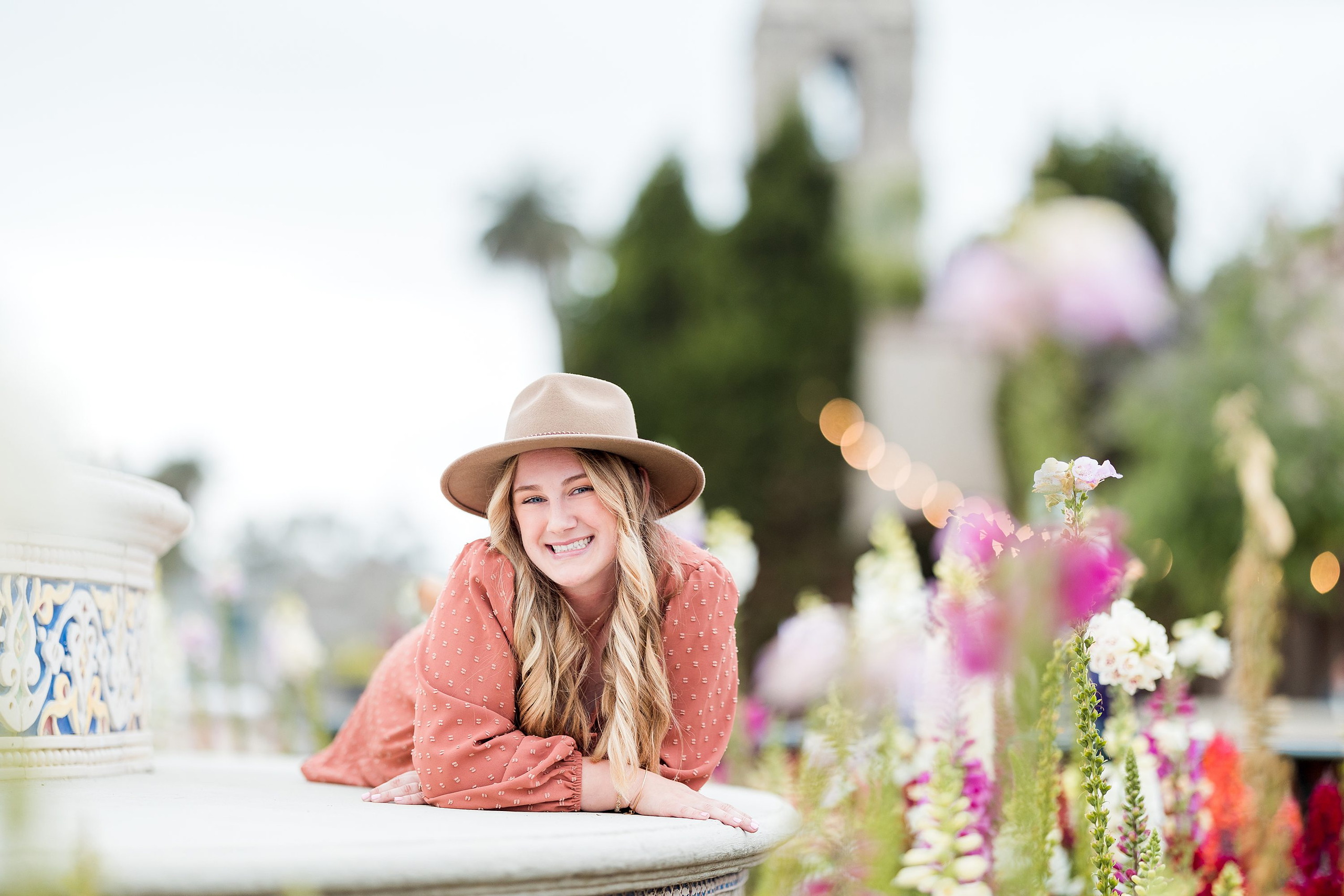 Balboa Park Senior Photography Session — 07 Senior leaning on white stone planter, pink dress and hat, bell tower visible