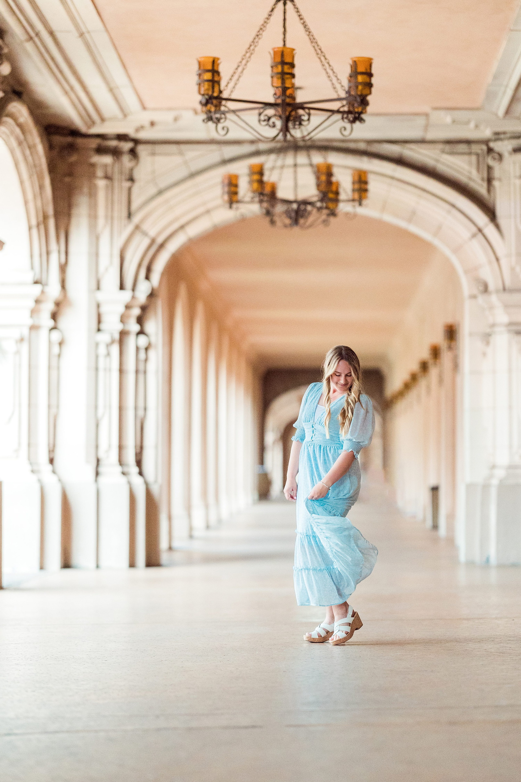 Balboa Park Senior Photography Session — 15 Senior in light blue dress on steps with arms spread wide near arched colonnade