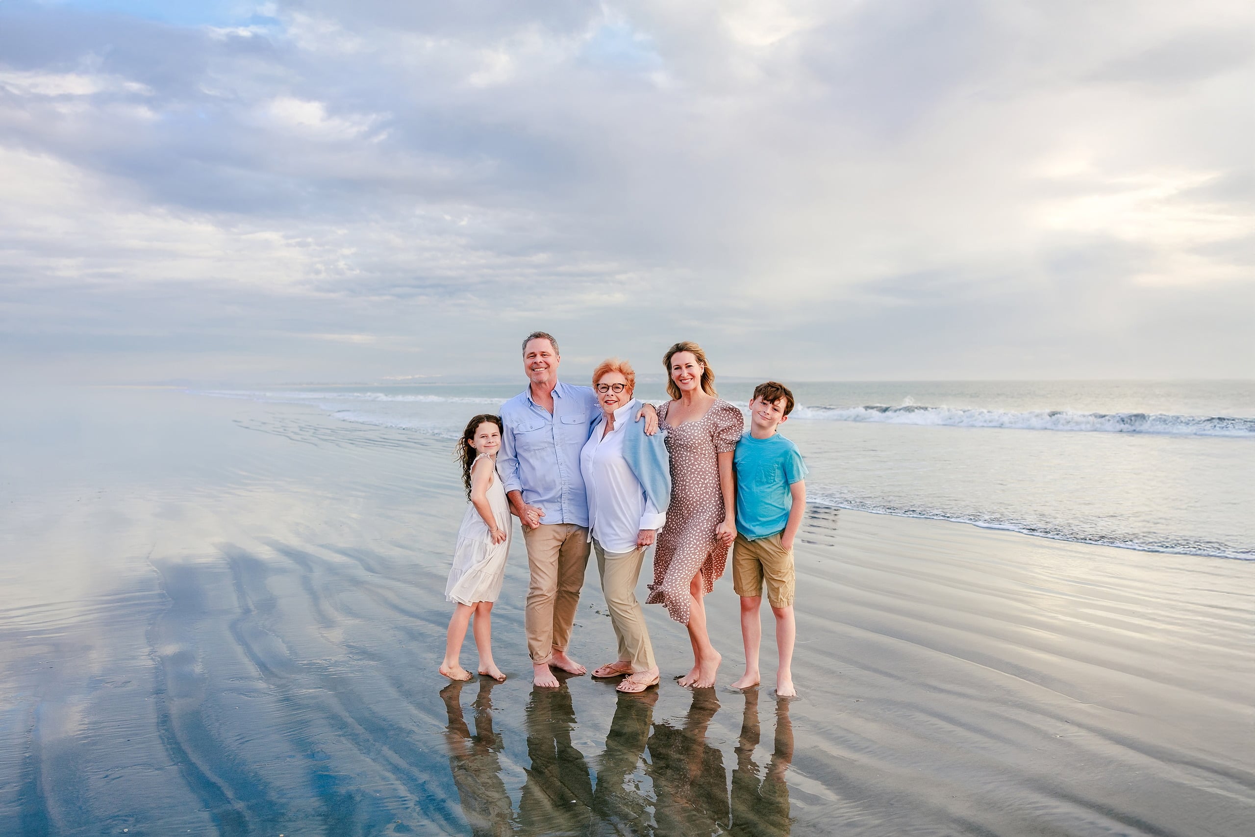 Behind the Scenes of Grammy Judy’s 80th Birthday Session: An Interview with Wendy — 01 Grammy Judy and family stand in shallow ocean water on beach under cloudy sky