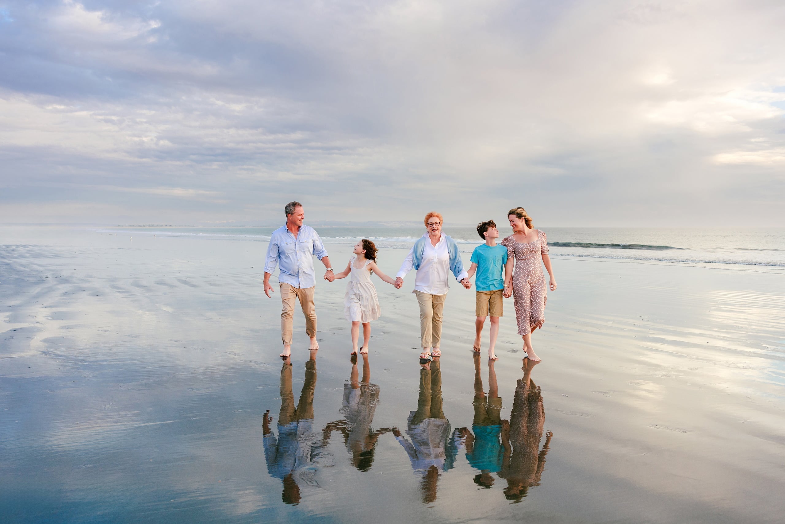 Behind the Scenes of Grammy Judy’s 80th Birthday Session: An Interview with Wendy — 02 Family of five walk through shallow water with perfect reflections on wet sand below