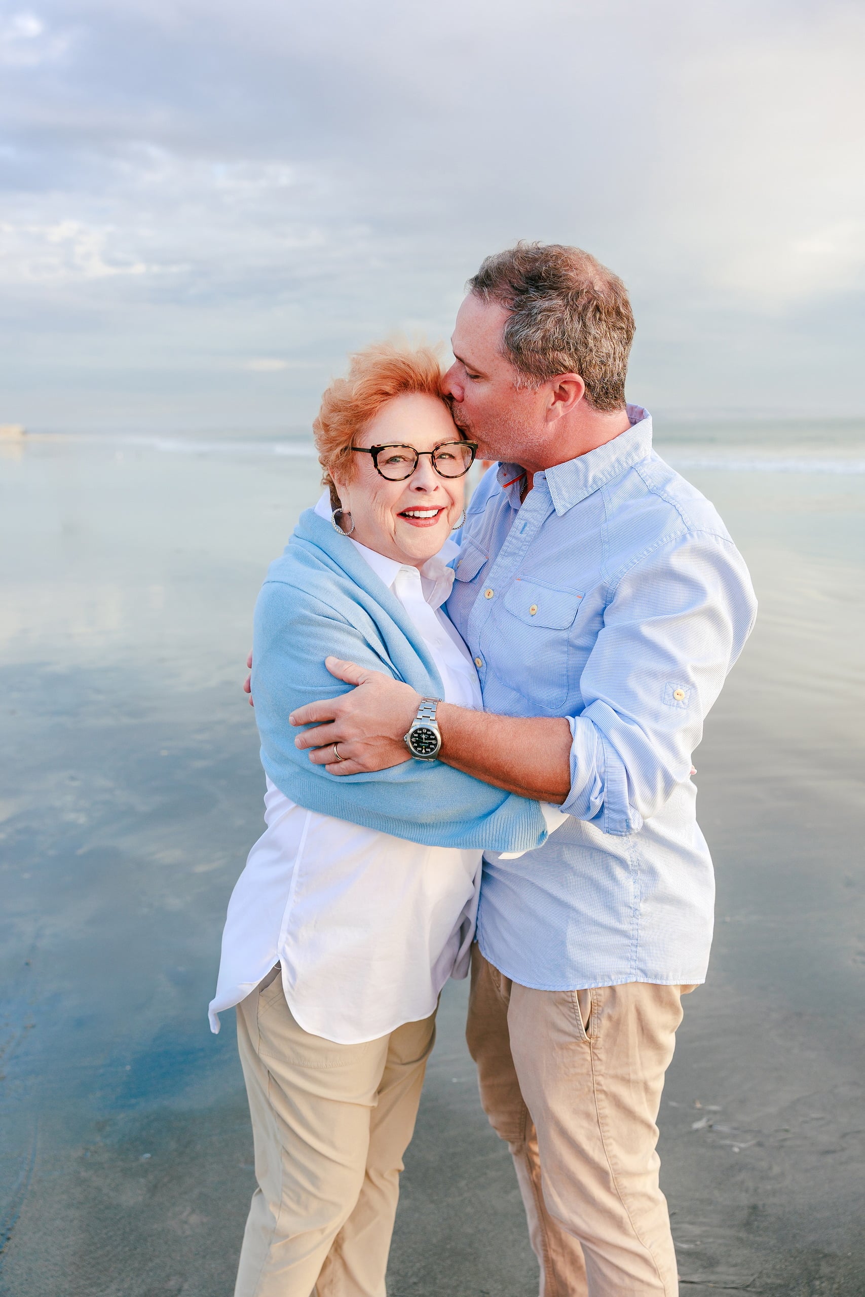 Behind the Scenes of Grammy Judy’s 80th Birthday Session: An Interview with Wendy — 07 Grammy Judy and man embrace on beach, both smiling, with ocean and sky behind them