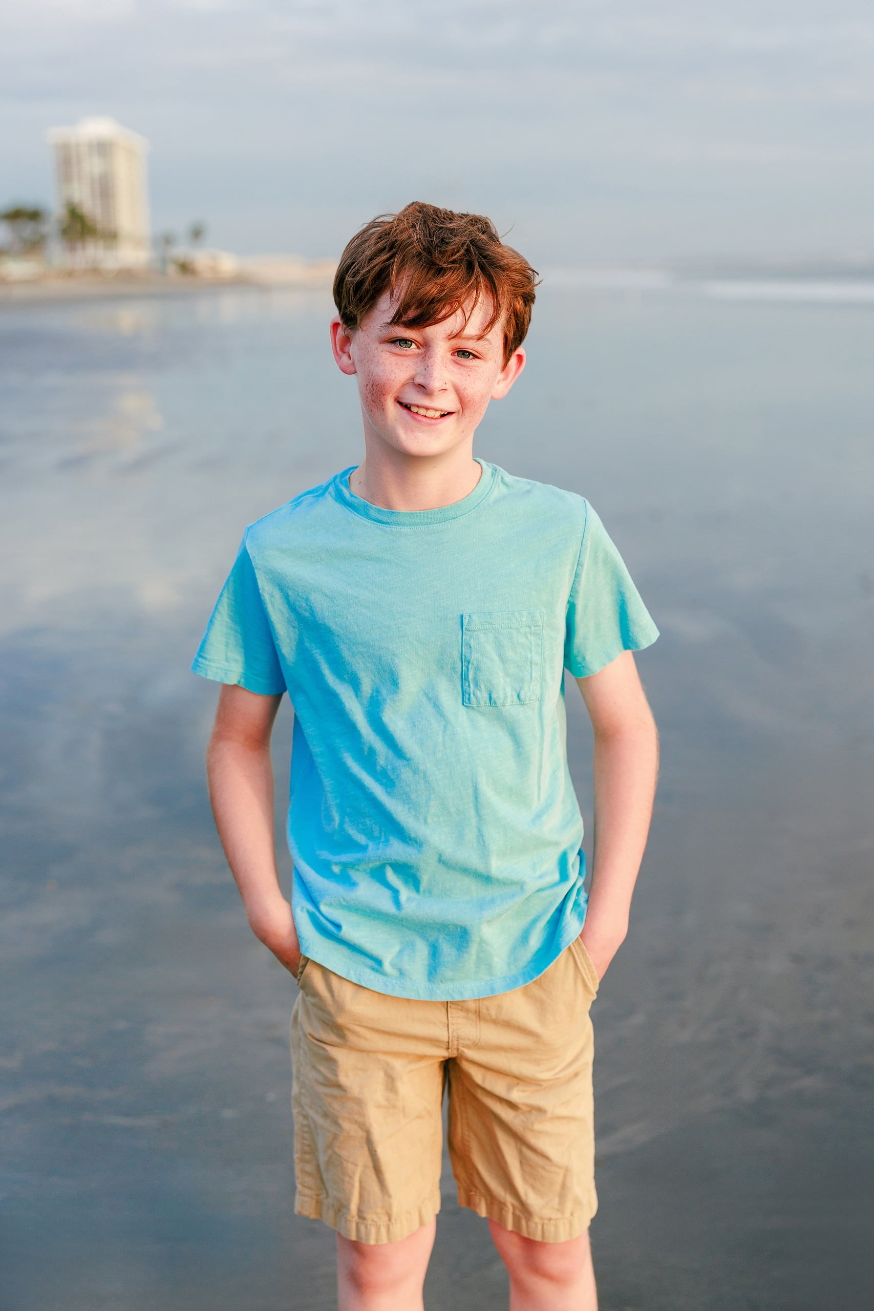 Behind the Scenes of Grammy Judy’s 80th Birthday Session: An Interview with Wendy — 09 Boy in turquoise shirt stands on beach with hands in pockets, Hotel del Coronado behind