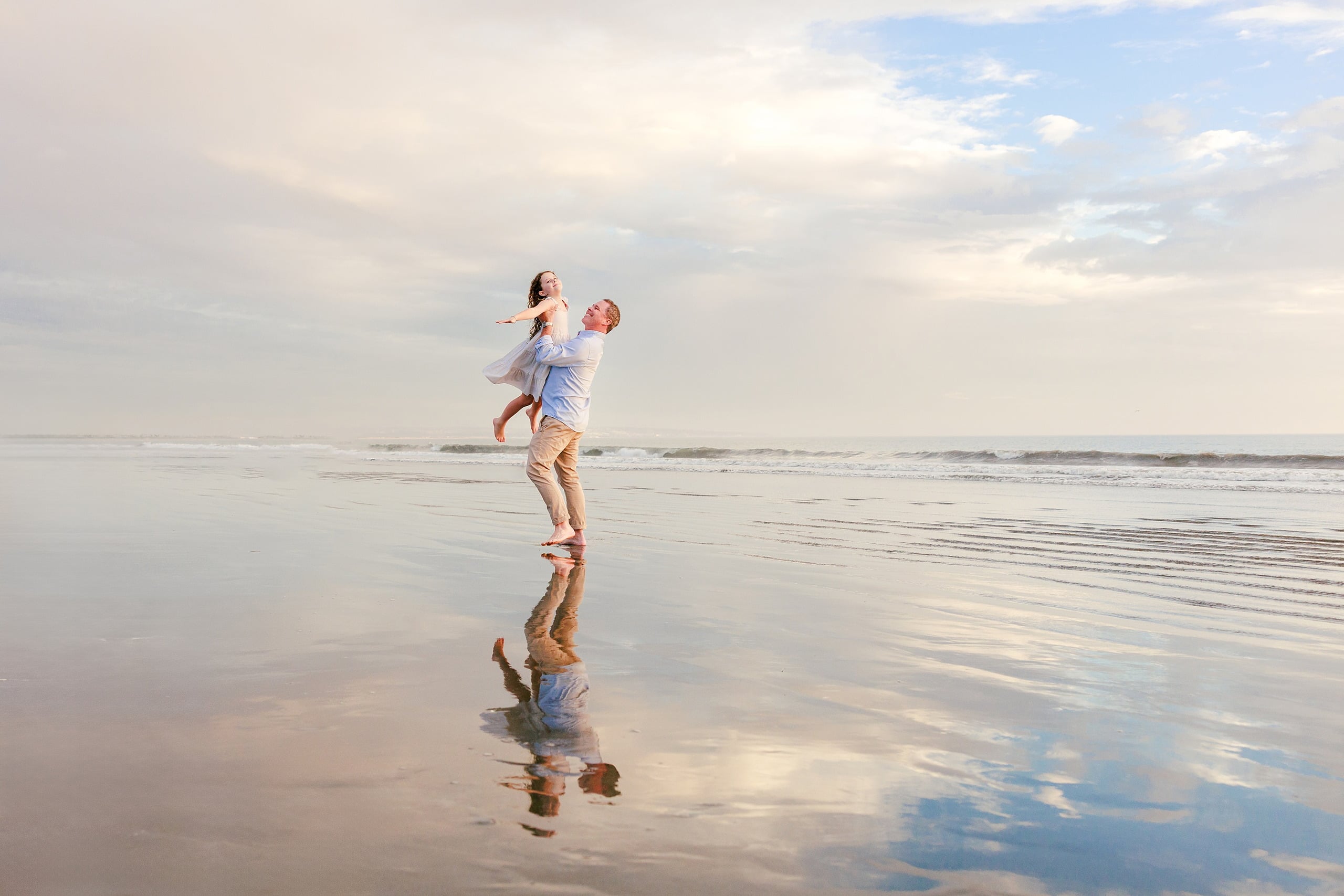 Behind the Scenes of Grammy Judy’s 80th Birthday Session: An Interview with Wendy — 11 Man lifts young girl laughing on beach at golden hour with dramatic sky and water reflections