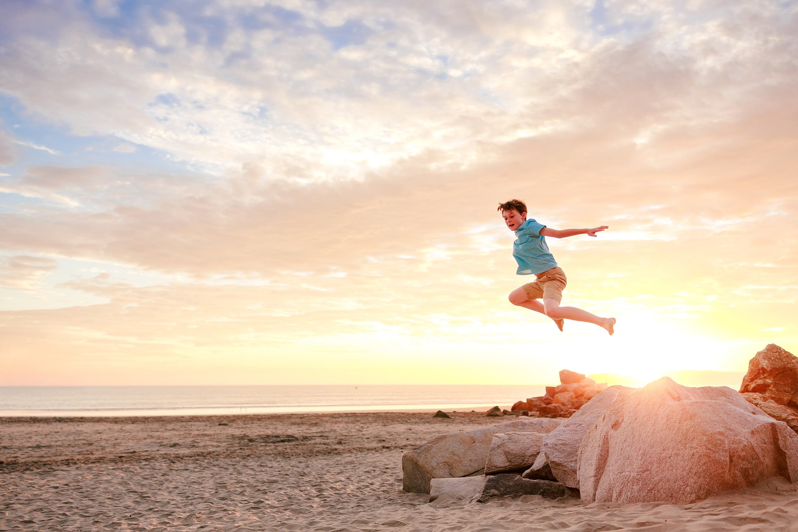 Behind the Scenes of Grammy Judy’s 80th Birthday Session: An Interview with Wendy — 19 Boy jumps off large rock at beach, framed against warm golden sunset light