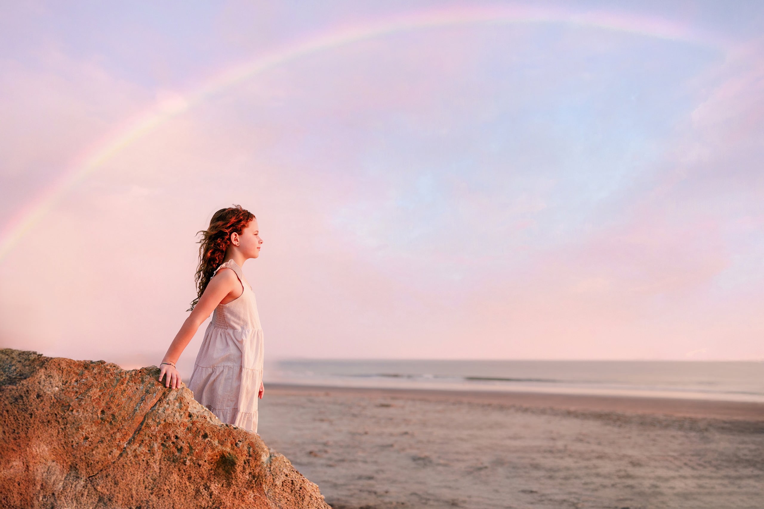 Behind the Scenes of Grammy Judy’s 80th Birthday Session: An Interview with Wendy — 20 Girl in white stands on rock at beach, silhouetted against sky with bright rainbow above