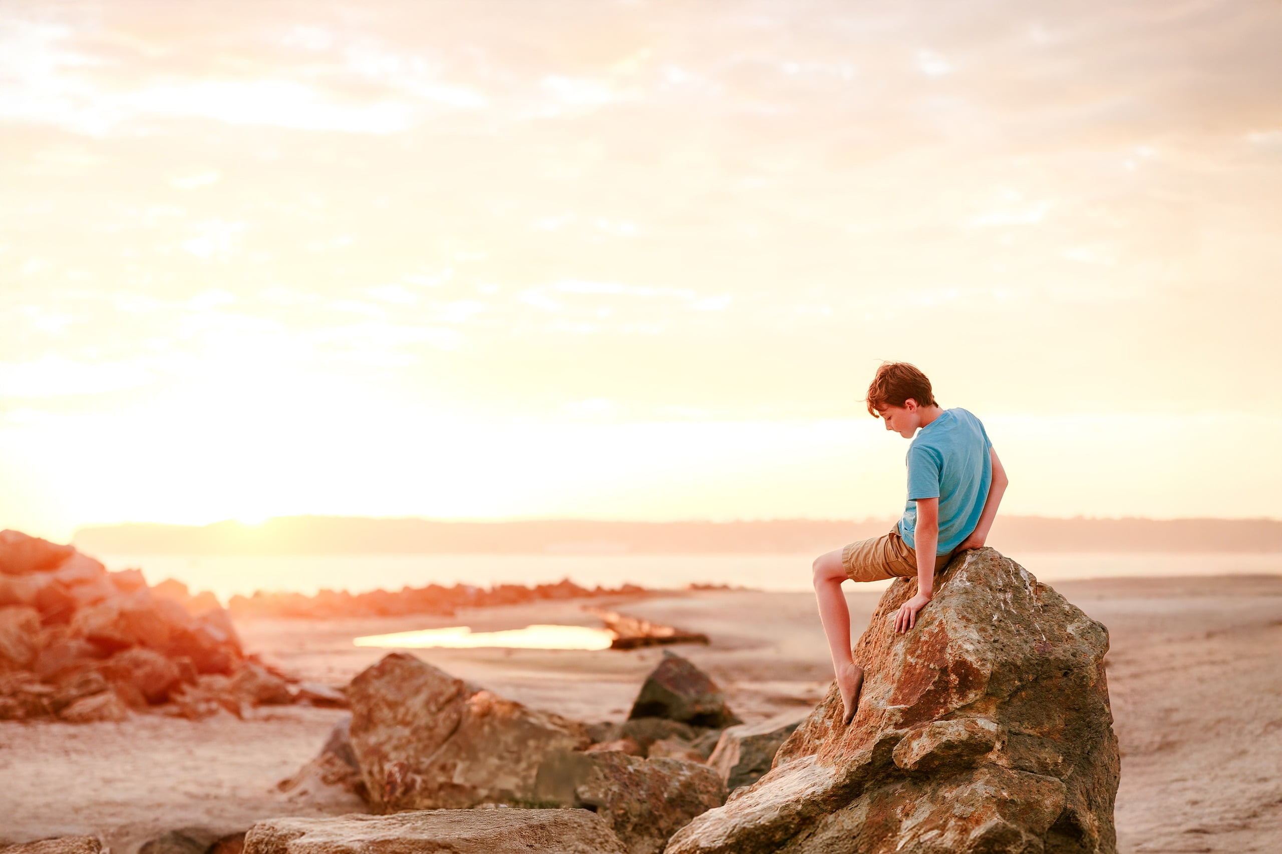 Behind the Scenes of Grammy Judy’s 80th Birthday Session: An Interview with Wendy — 23 Boy sits on large rock at beach at golden hour, looking down as sun sets