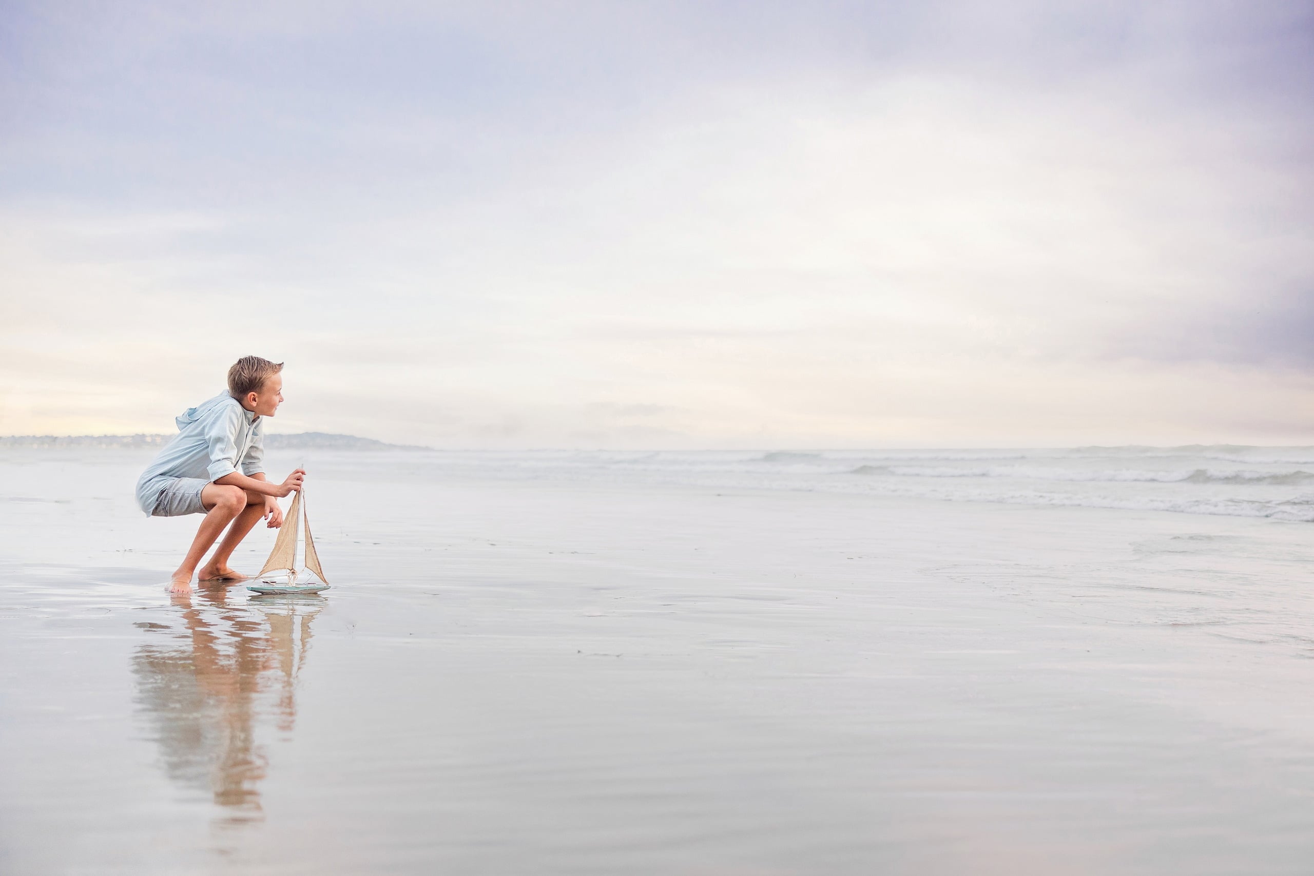 Butlers on the Beach — 19 Boy crouching down to hold small sailboat toy in shallow water