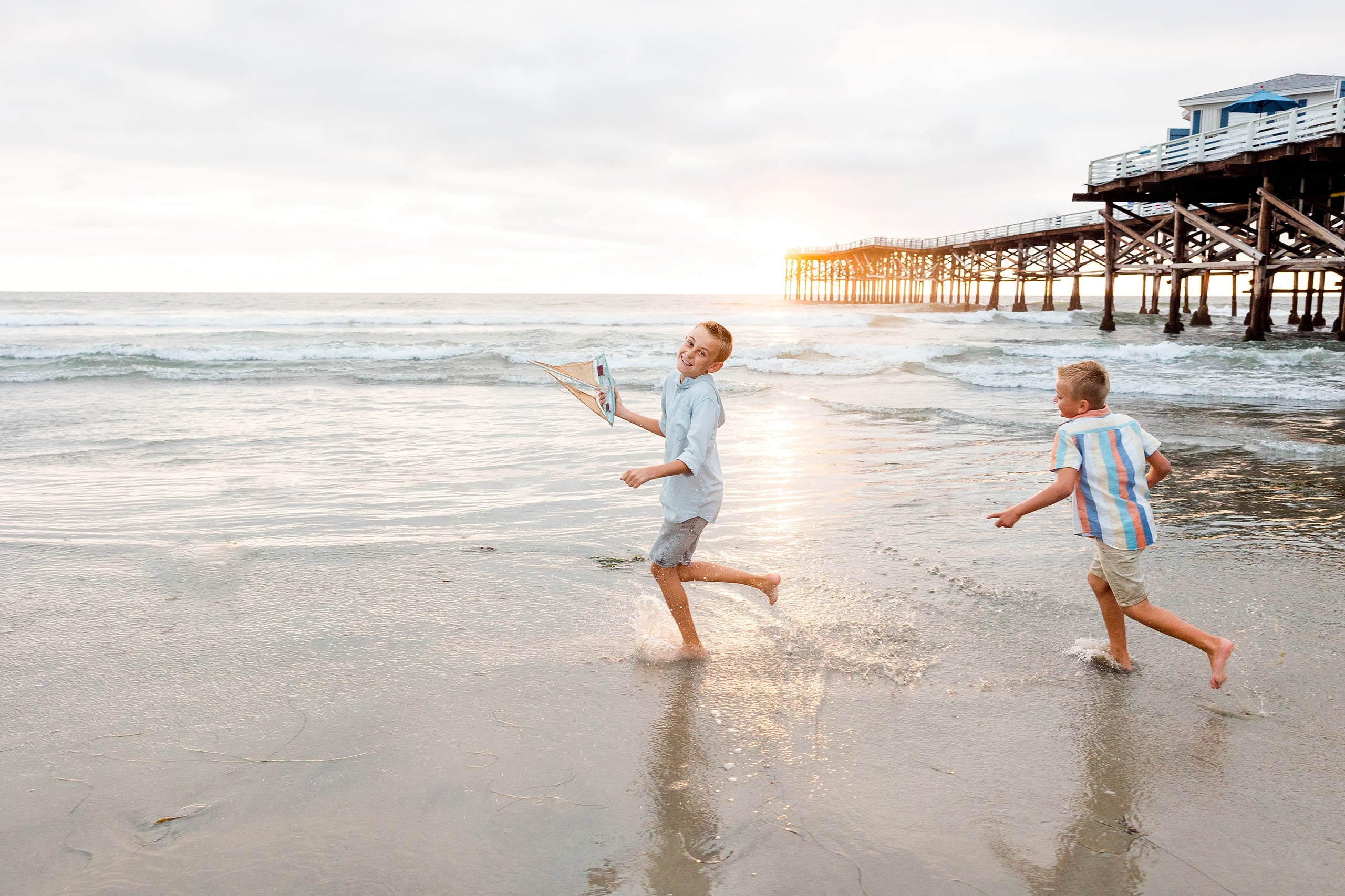 Butlers on the Beach — 21 Two boys racing through shallow water each holding toy sailboats at pier