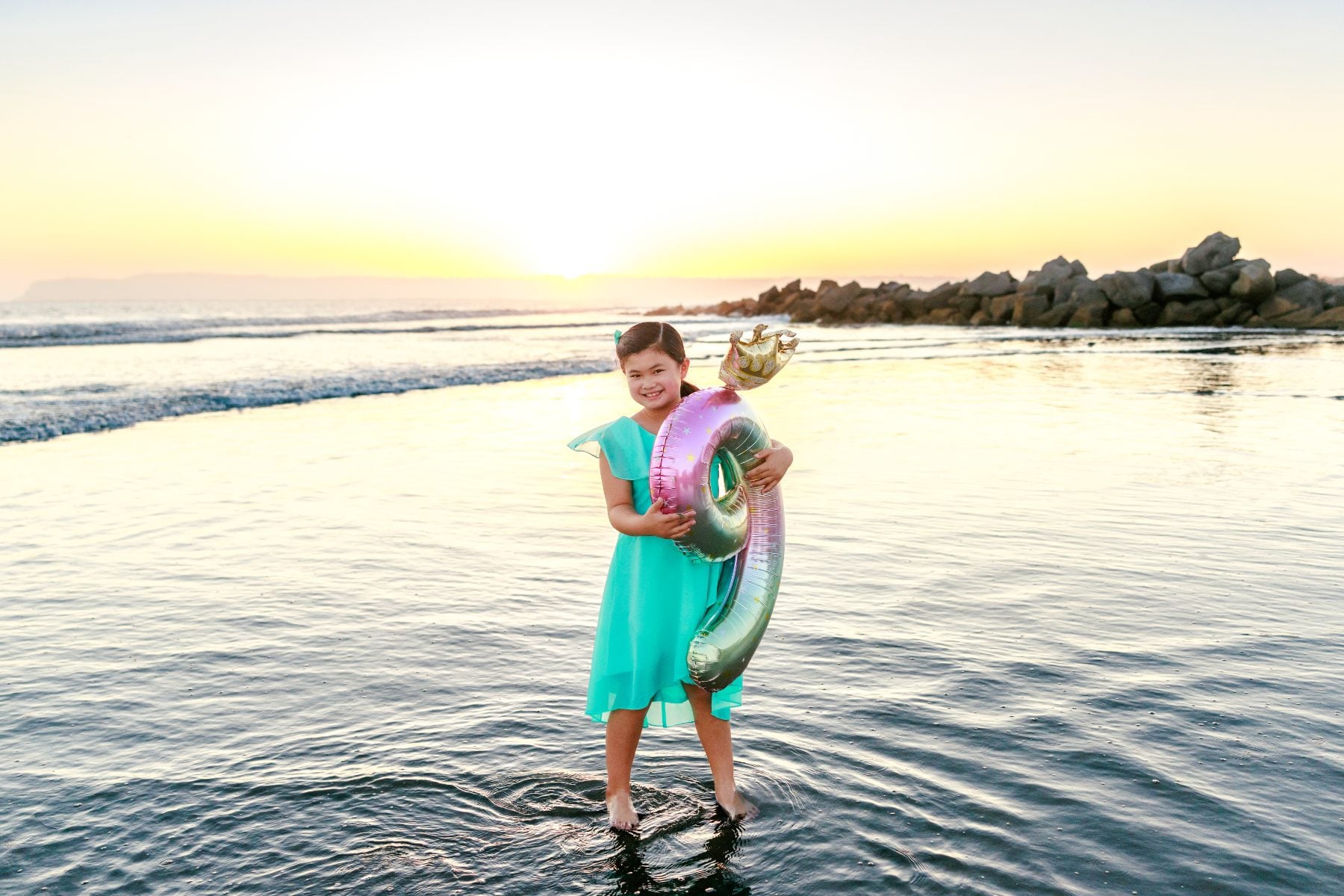 Girl holding mermaid balloon wading in shallow water at Coronado Beach sunset
