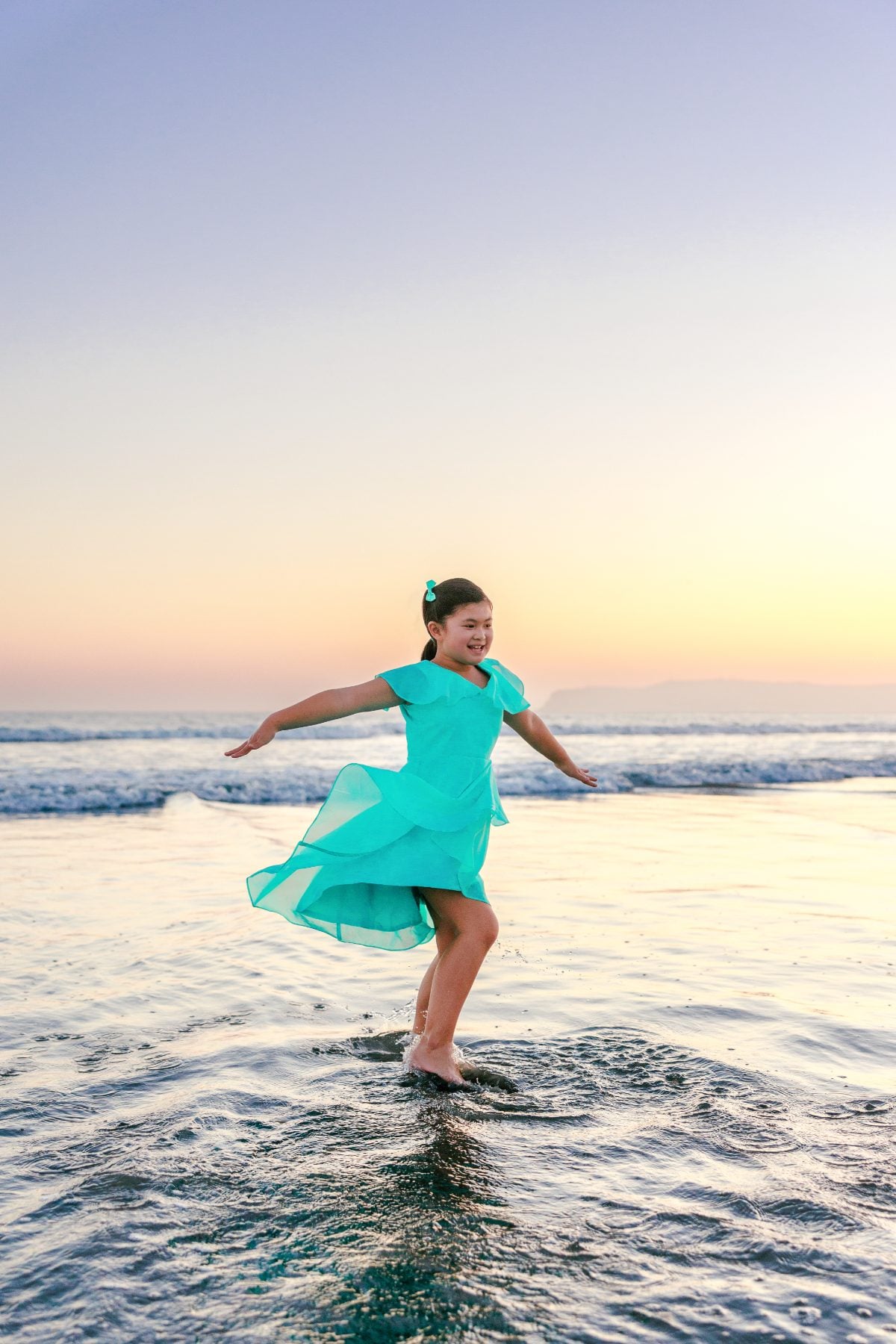 Girl in teal dress twirling barefoot in ocean waves at golden hour