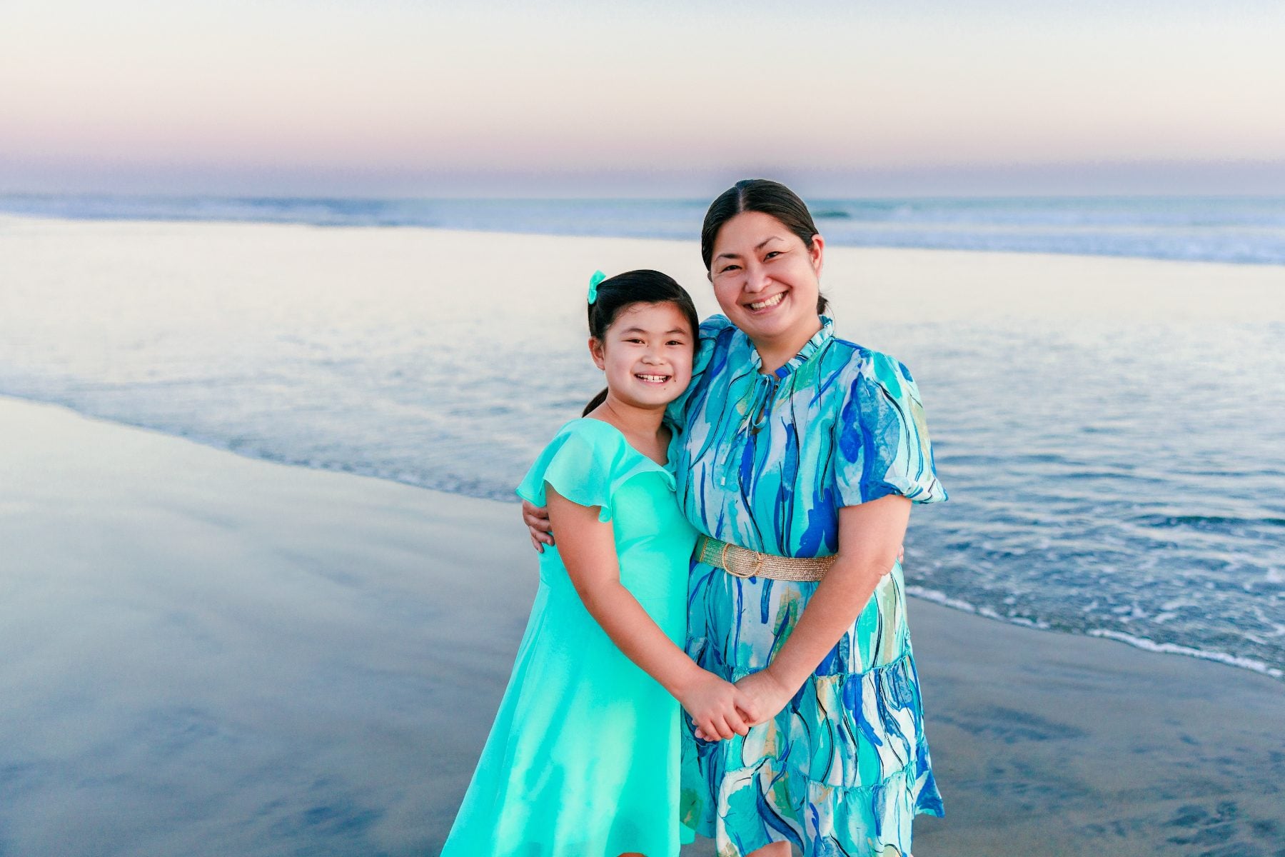 Mother and daughter smiling together on the sand as waves roll in at sunset