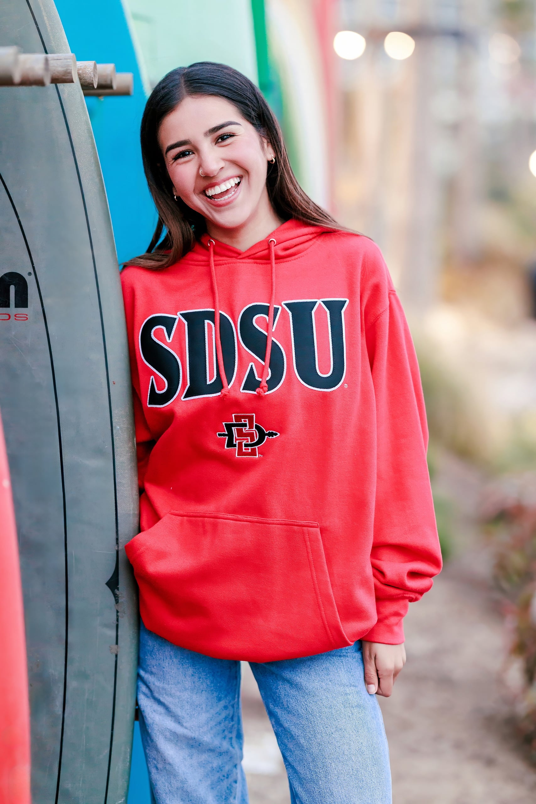 Evanna, Atalia, and Erleen’s Senior Session on Coronado — 37 Senior in red SDSU hoodie standing by colorful surfboards at beach