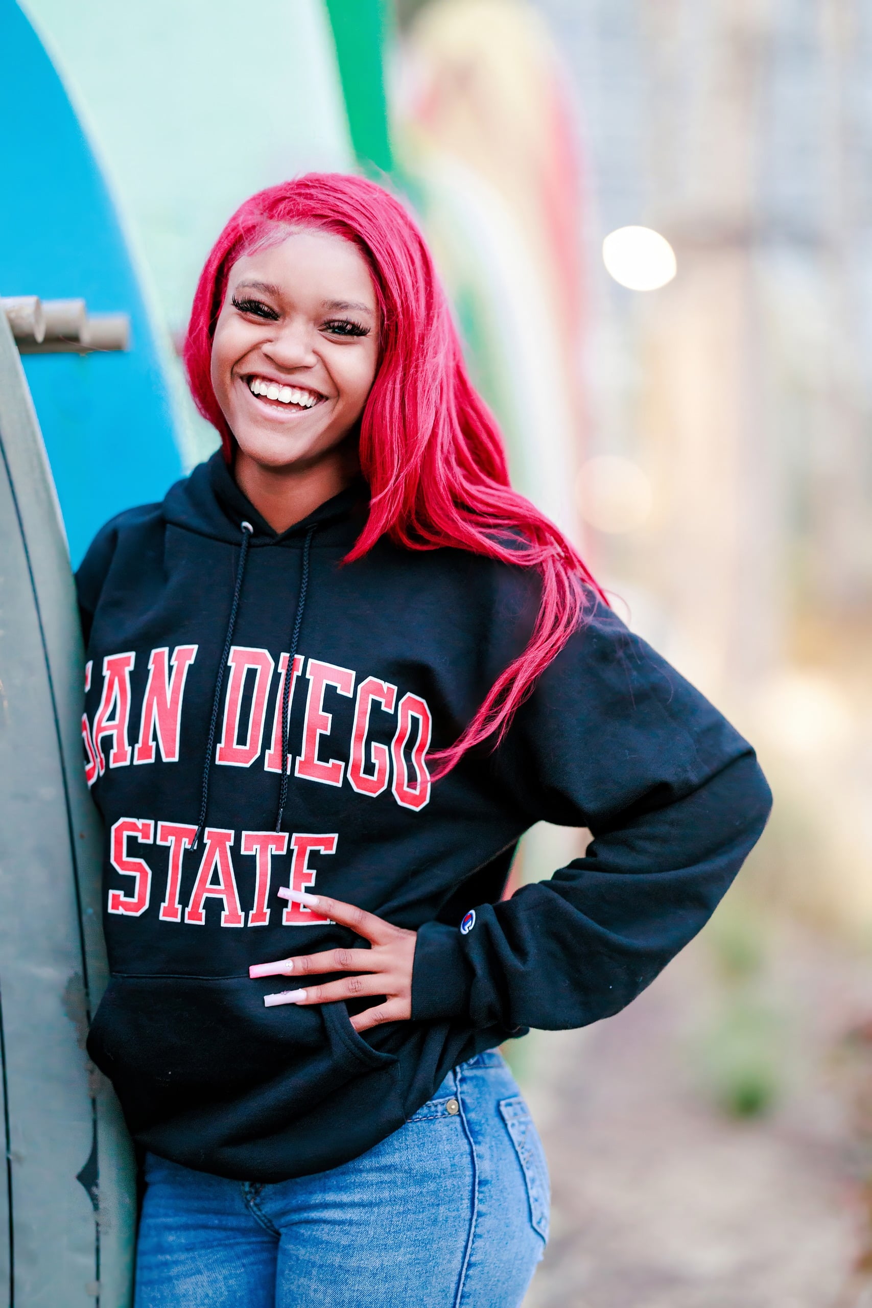 Evanna, Atalia, and Erleen’s Senior Session on Coronado — 38 Senior with pink hair in San Diego State hoodie by painted surfboards
