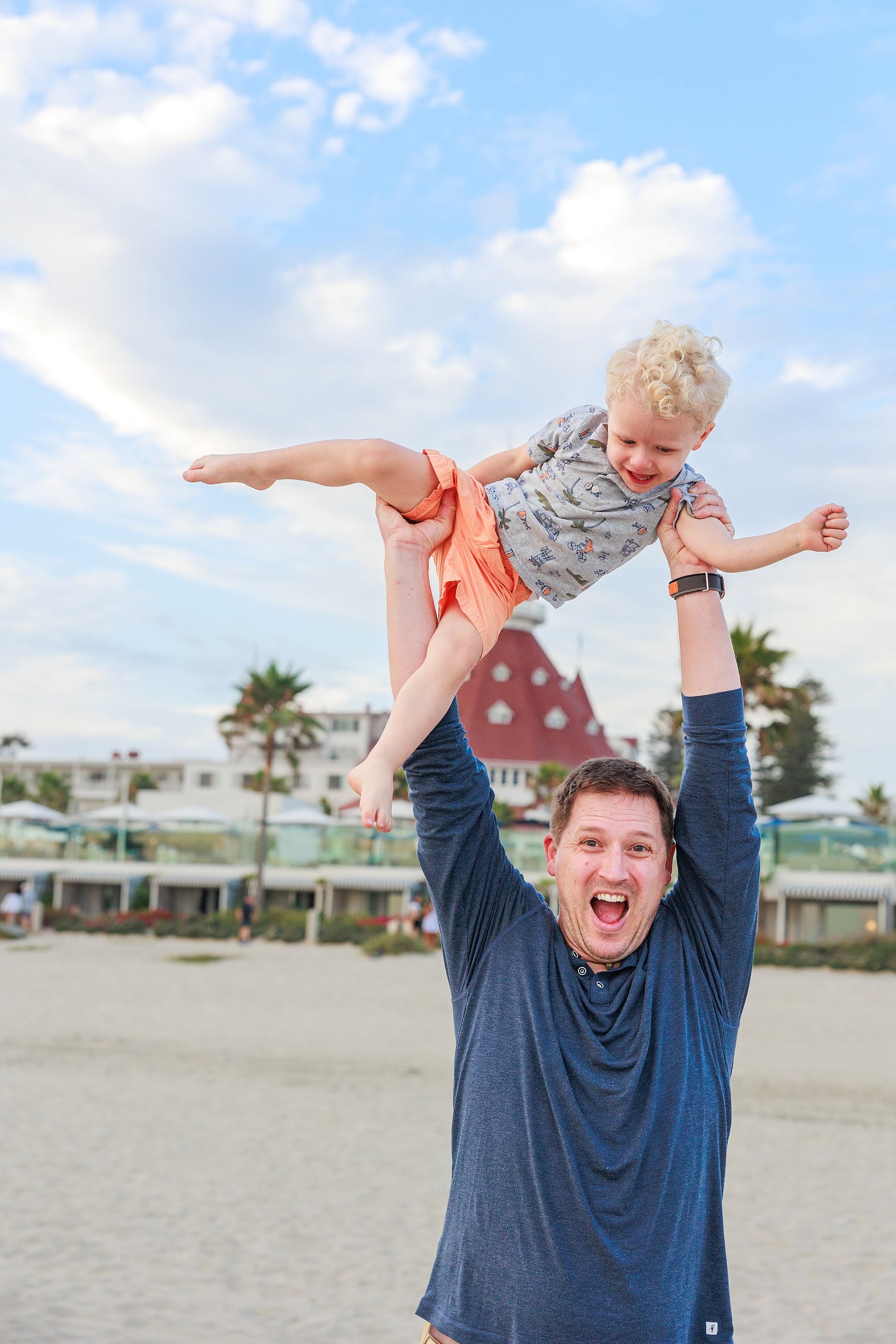 Extended Family Photography Hotel Del Coronado — 06 Adult man hoists small child overhead on beach, Hotel del Coronado beyond