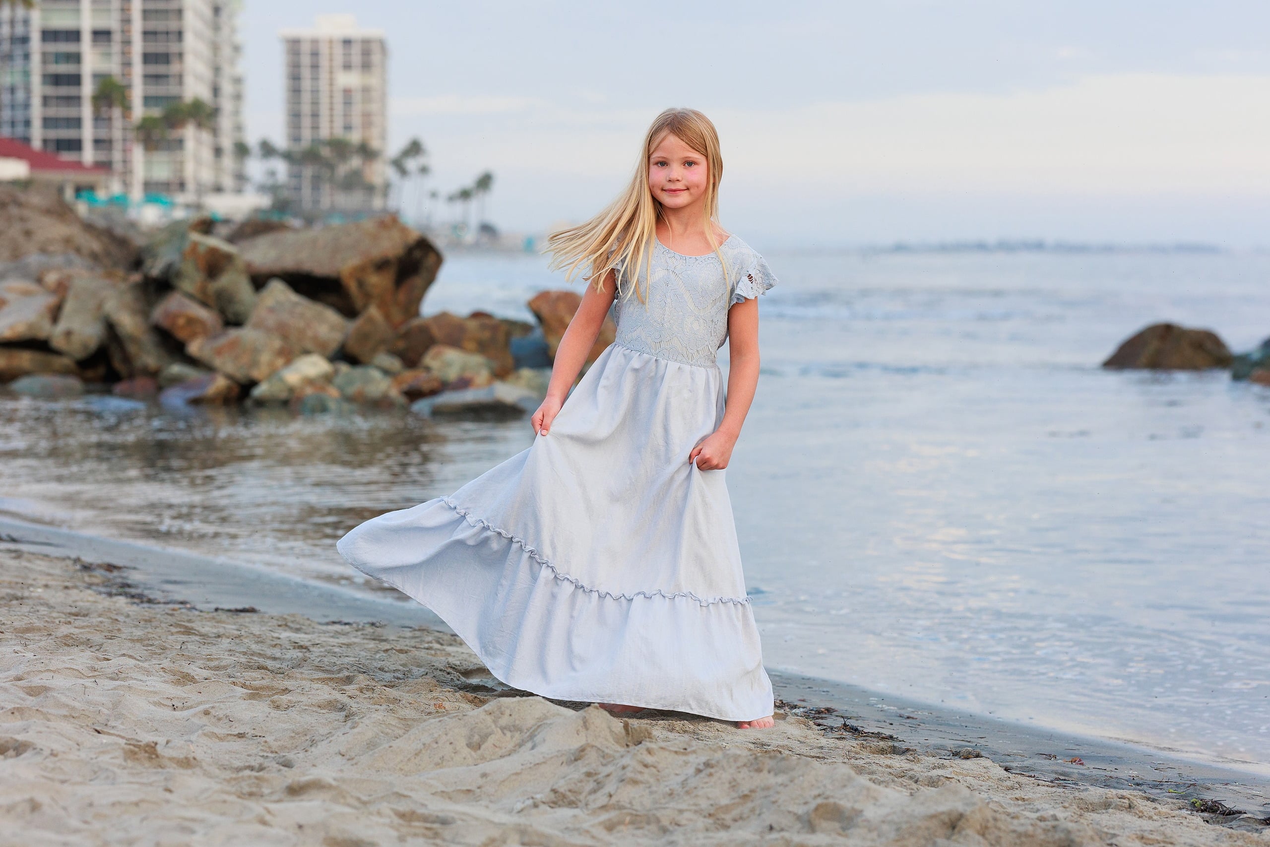 Extended Family Photography Hotel Del Coronado — 19 Child in white dress walks along shoreline with coastal city buildings behind