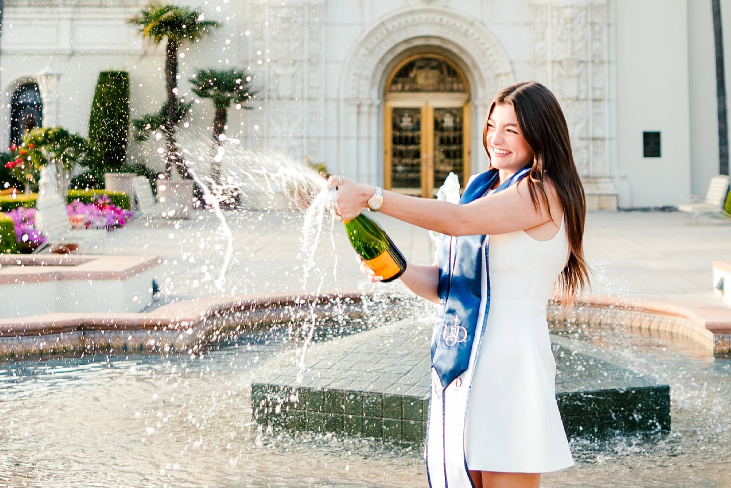 Kenni’s USD grad Session — 05 Kenni celebrates at fountain in white dress and blue USD sash with champagne bottle