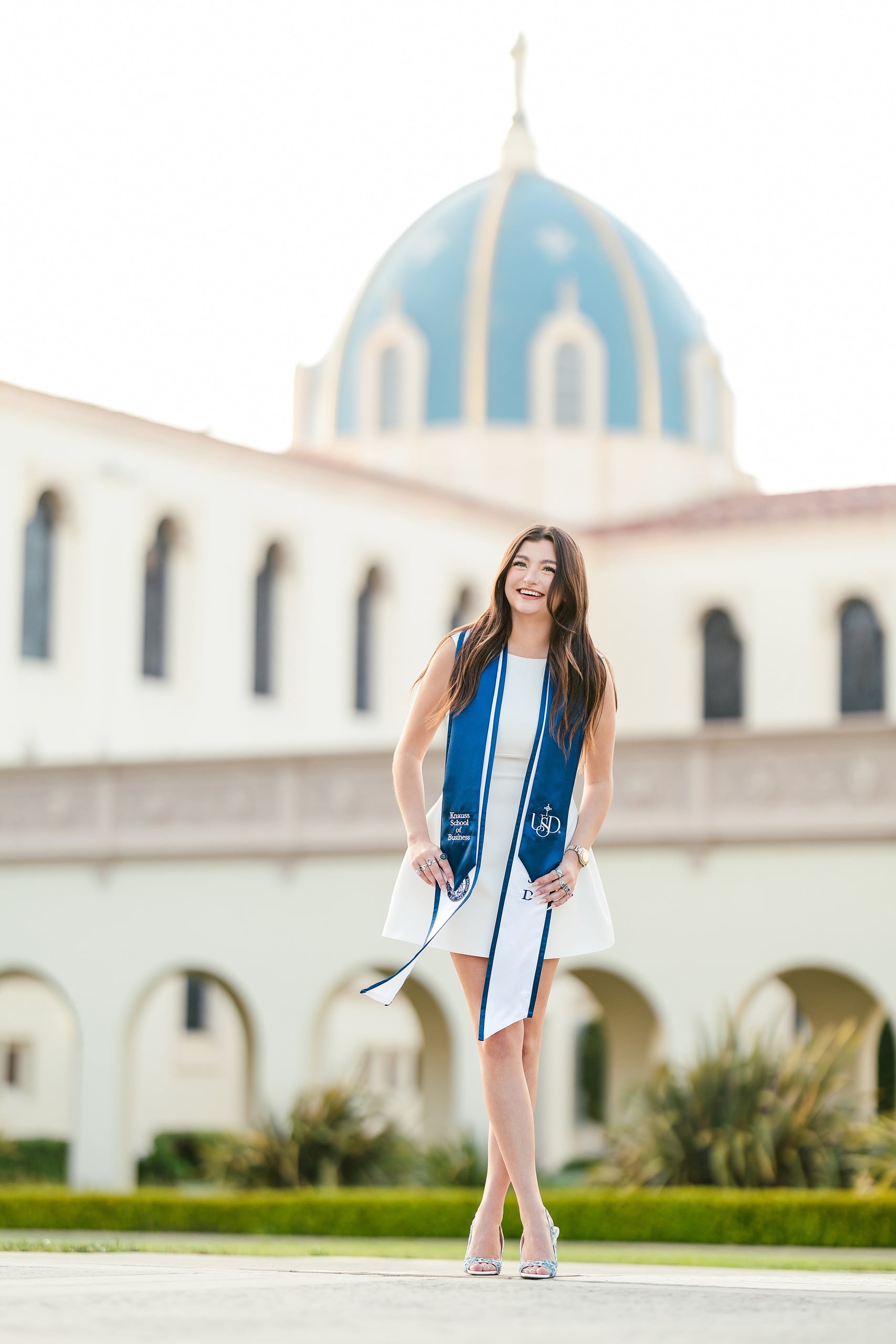 Kenni’s USD grad Session — 11 Kenni smiles in front of Immaculata at USD wearing white dress and blue sash