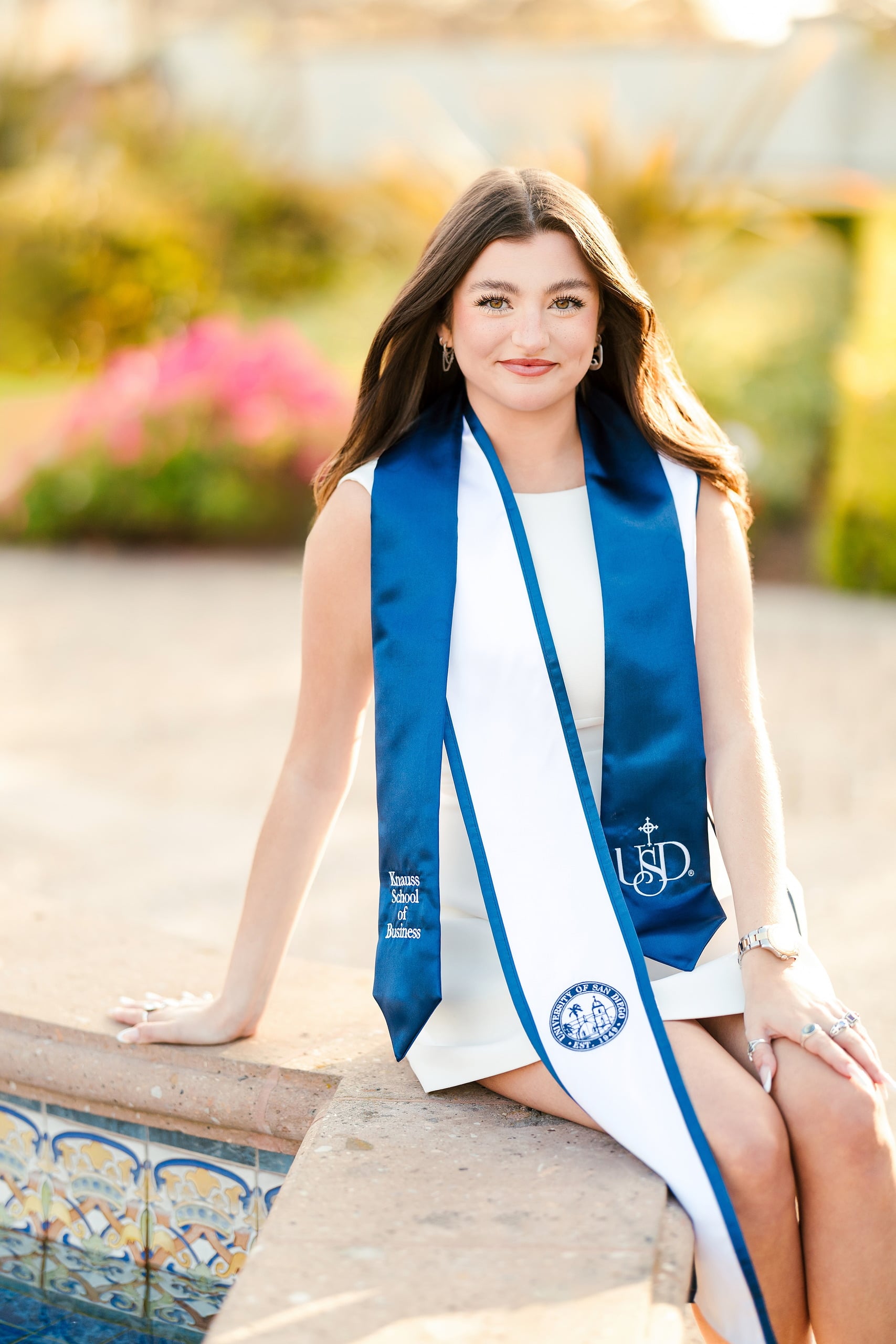 Kenni’s USD grad Session — 12 Kenni sits on ornamental tile at USD courtyard in white dress with blue sash, golden hour