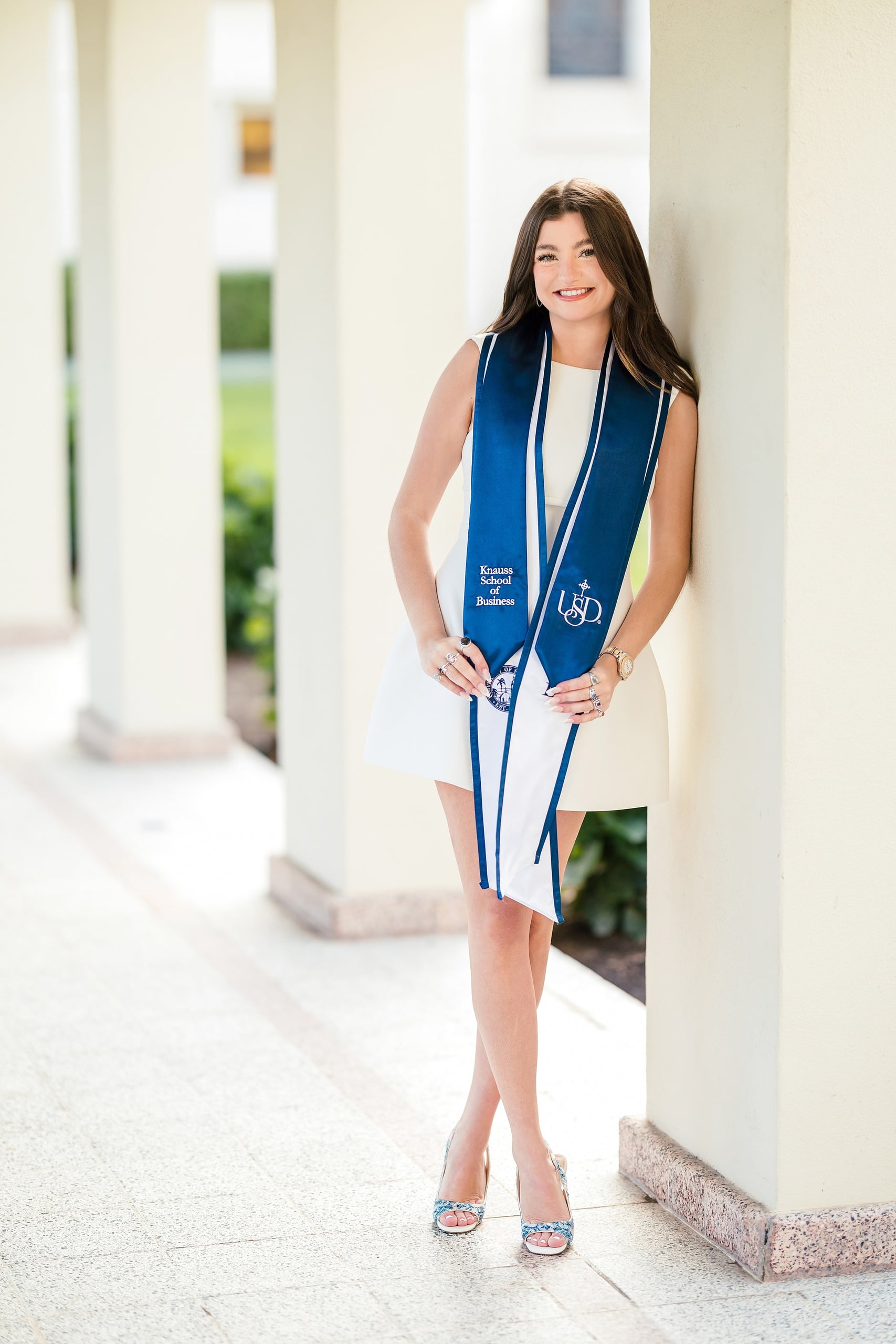 Kenni’s USD grad Session — 13 Kenni stands at colonnade at USD in white dress and blue sash, hands at sides
