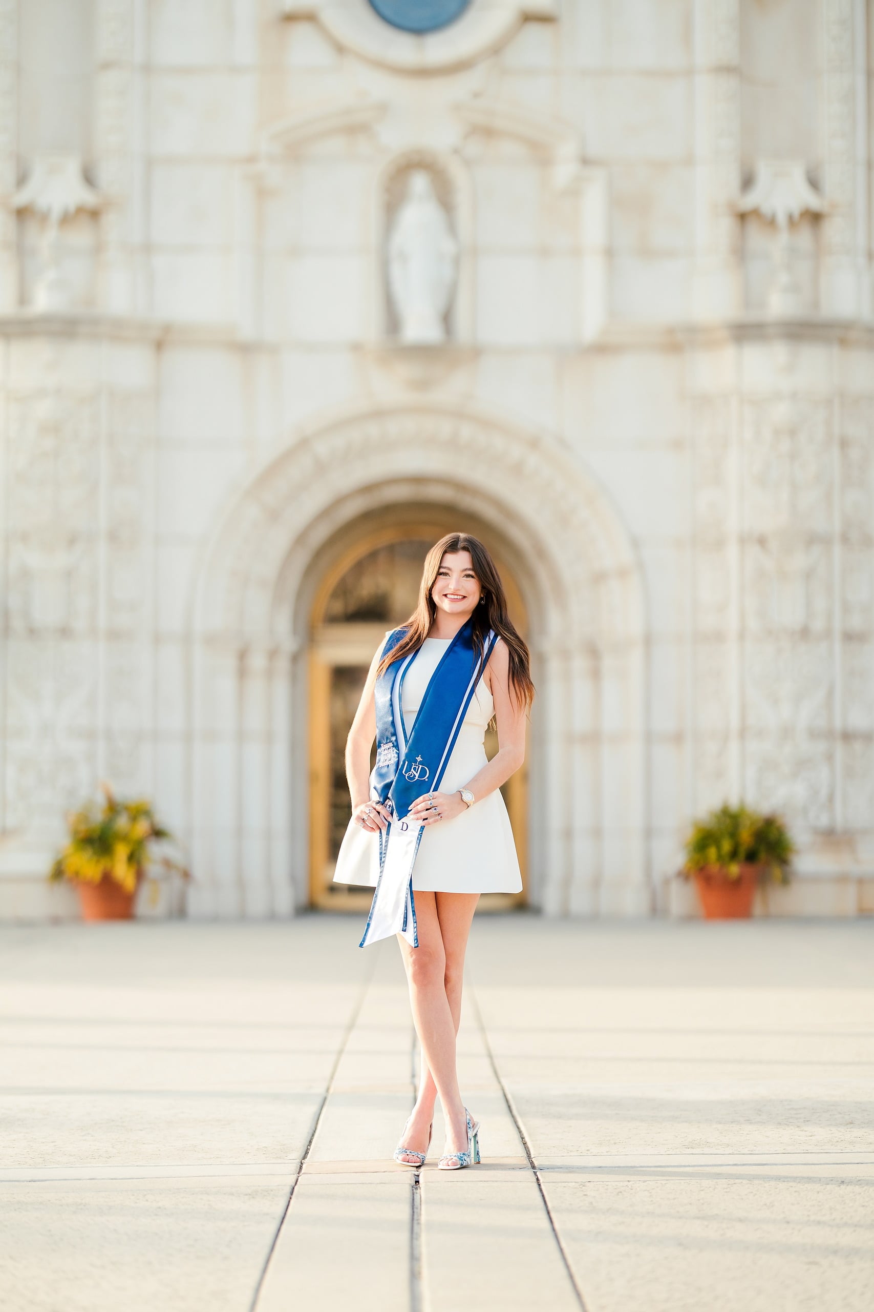 Kenni’s USD grad Session — 16 Kenni in white dress with blue sash before white mission church with golden doorway
