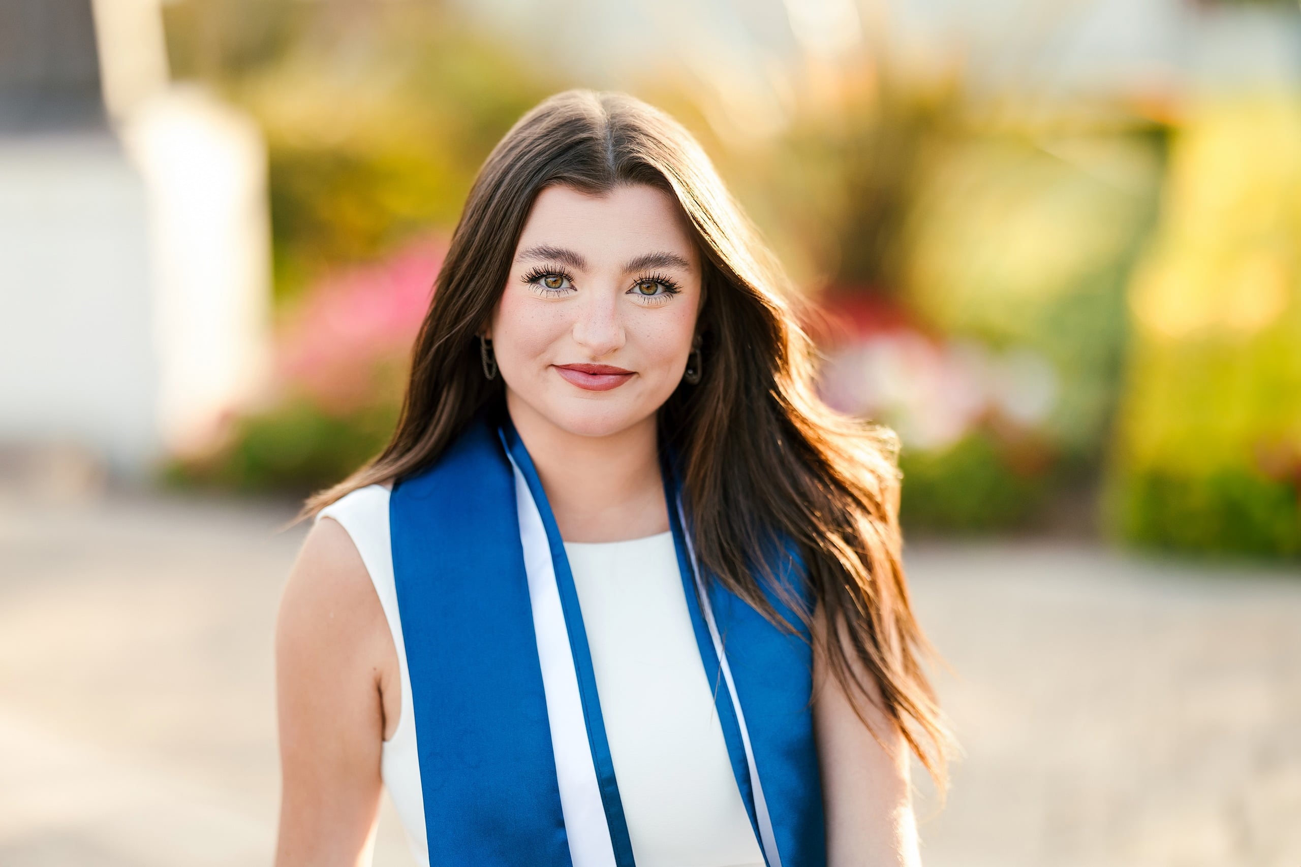 Kenni’s USD grad Session — 17 Kenni, close-up in white dress and blue sash with golden hour light, flowers softly blurred