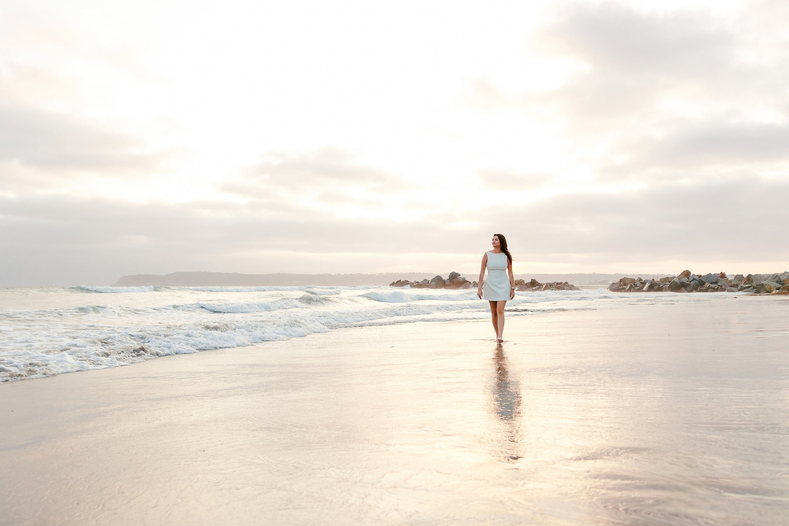 Kenni’s USD grad Session — 20 Kenni walks along beach in white dress with ocean waves and distant coastline visible