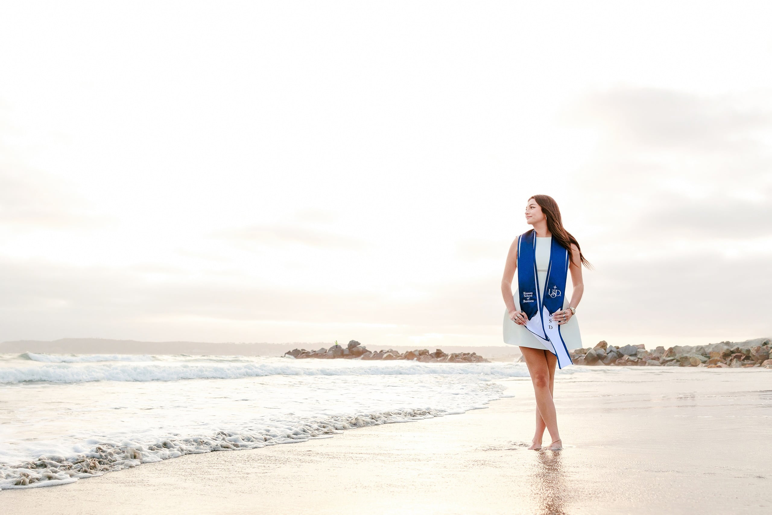 Kenni’s USD grad Session — 21 Kenni stands on beach in white dress with blue sash, gentle waves visible in background