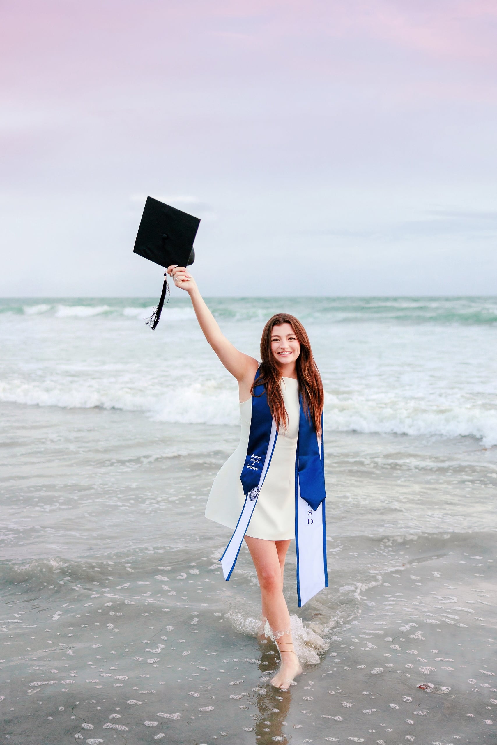 Kenni’s USD grad Session — 22 Kenni on beach at sunset in white dress with blue sash, graduation cap held overhead