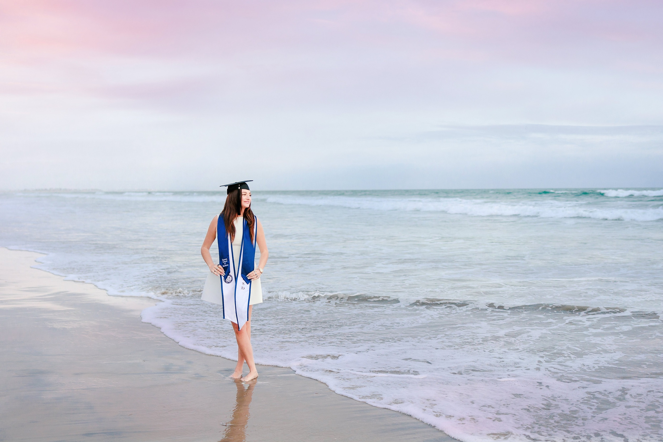 Kenni’s USD grad Session — 23 Kenni wades in shallow water on beach in white dress with blue sash at golden hour