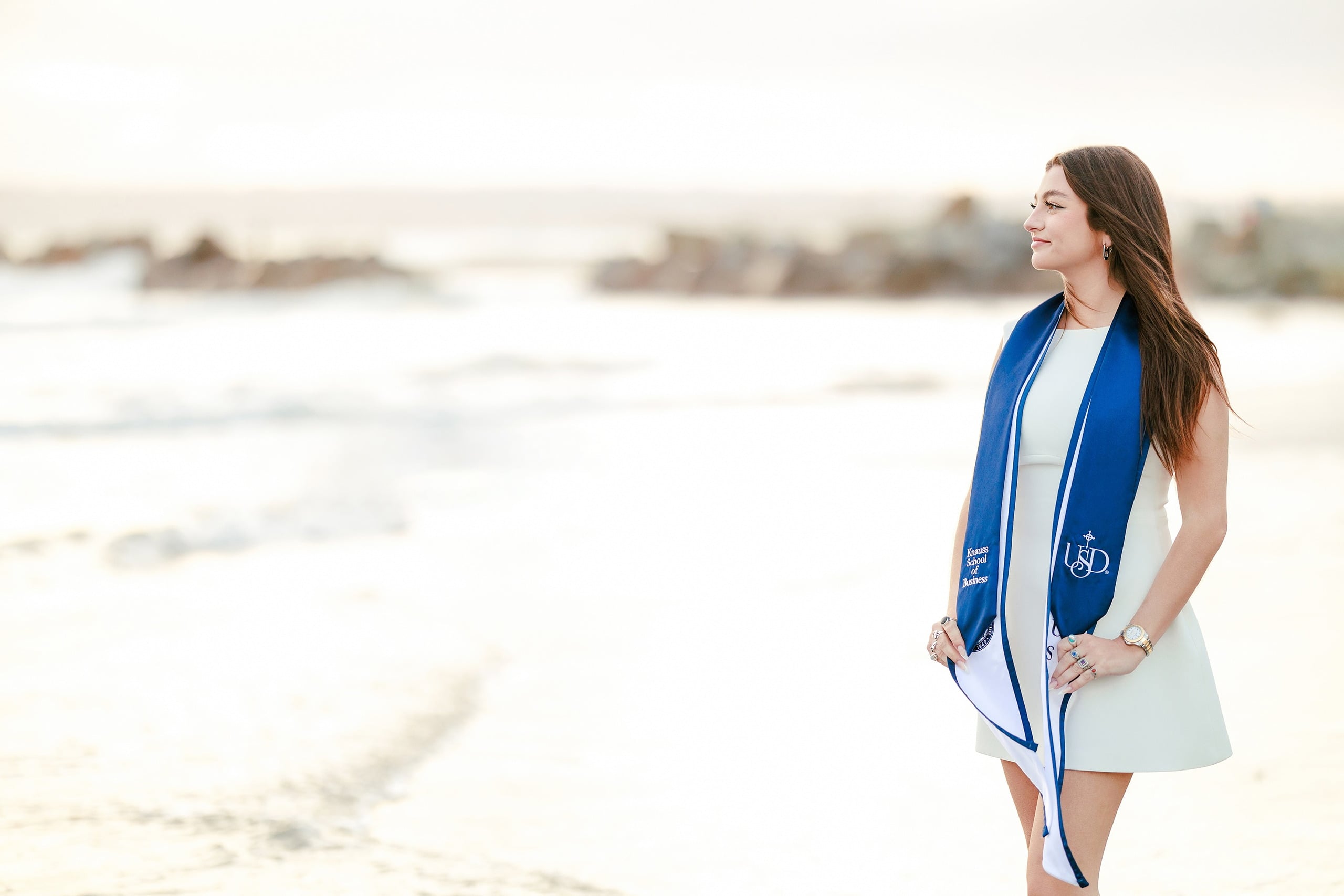 Kenni’s USD grad Session — 24 Kenni in sand wearing white dress with blue sash, gazing out toward calm ocean waves