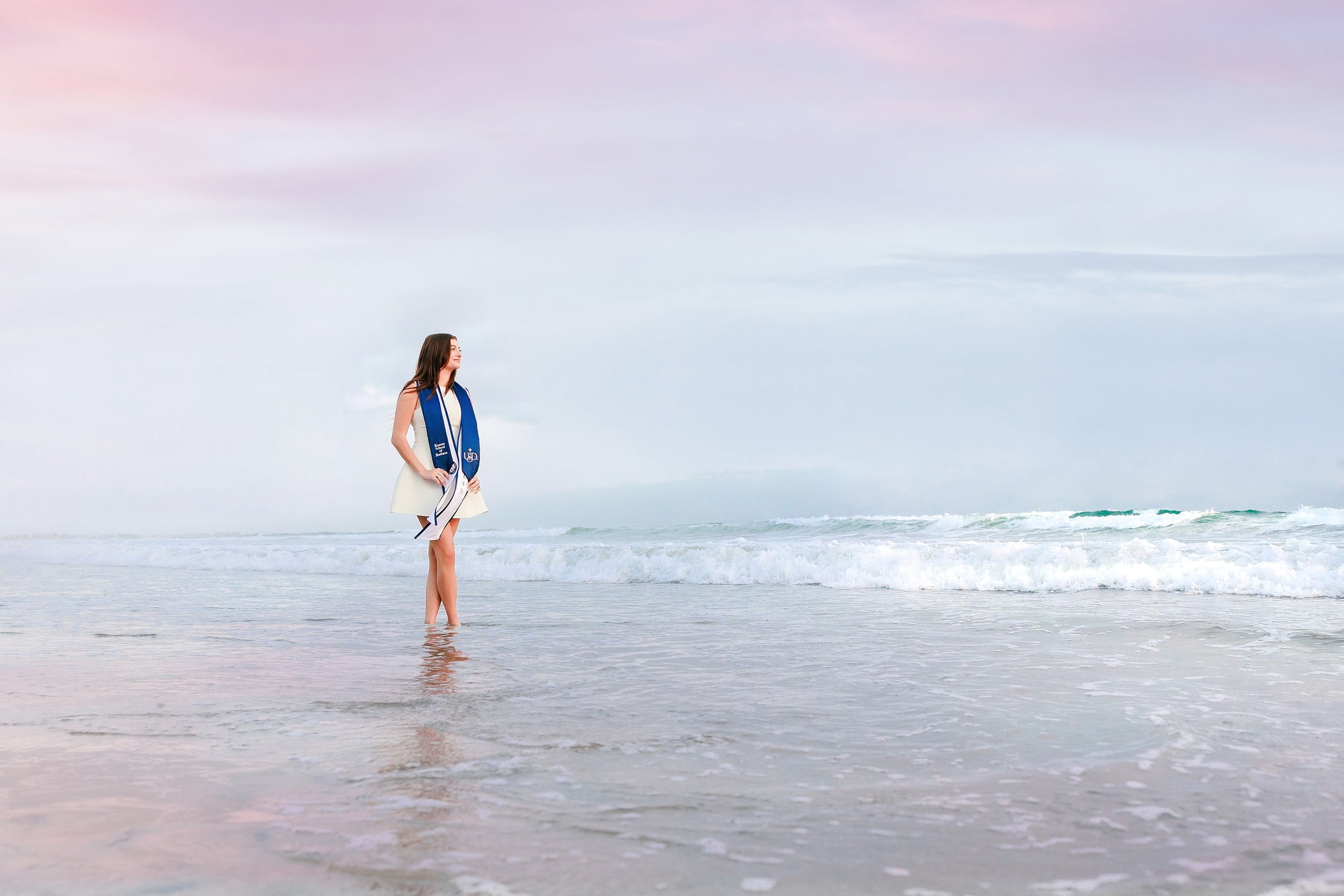 Kenni’s USD grad Session — 25 Kenni wades in shallow water on beach in white dress and blue sash at soft dusk light