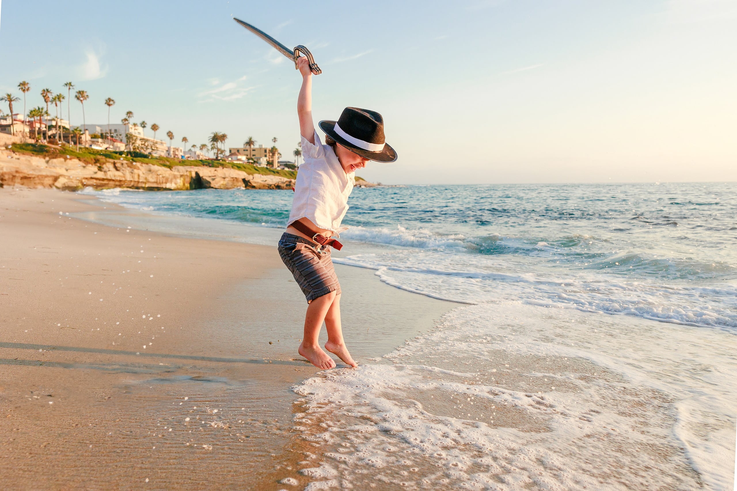 La Jolla Family Photography — 03 Child holding pirate sword raised high on La Jolla beach with palm tree bluff visible